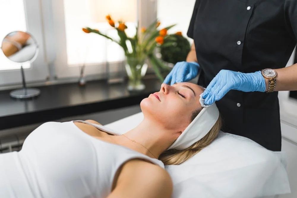 A woman receiving a facial treatment at a spa, lying on a treatment bed with a towel on her head, while a professional in black scrubs wearing blue gloves applies a facial mask or cream.