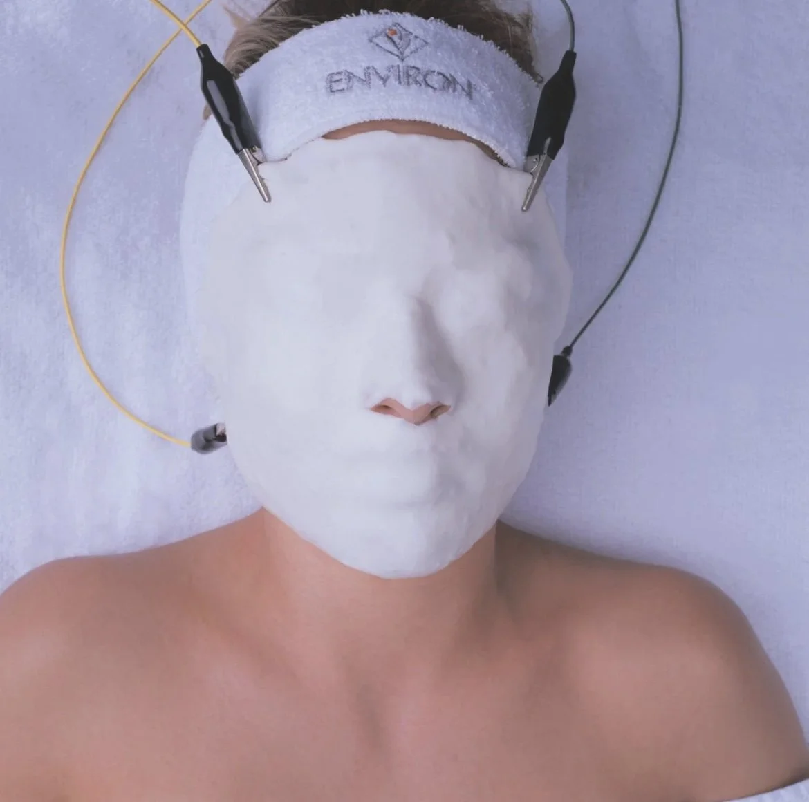 A person lying on a white surface with a white mask covering their face, connected to electrodes and wires, and wearing a headband with the word 'ENVIRON' embroidered on it.