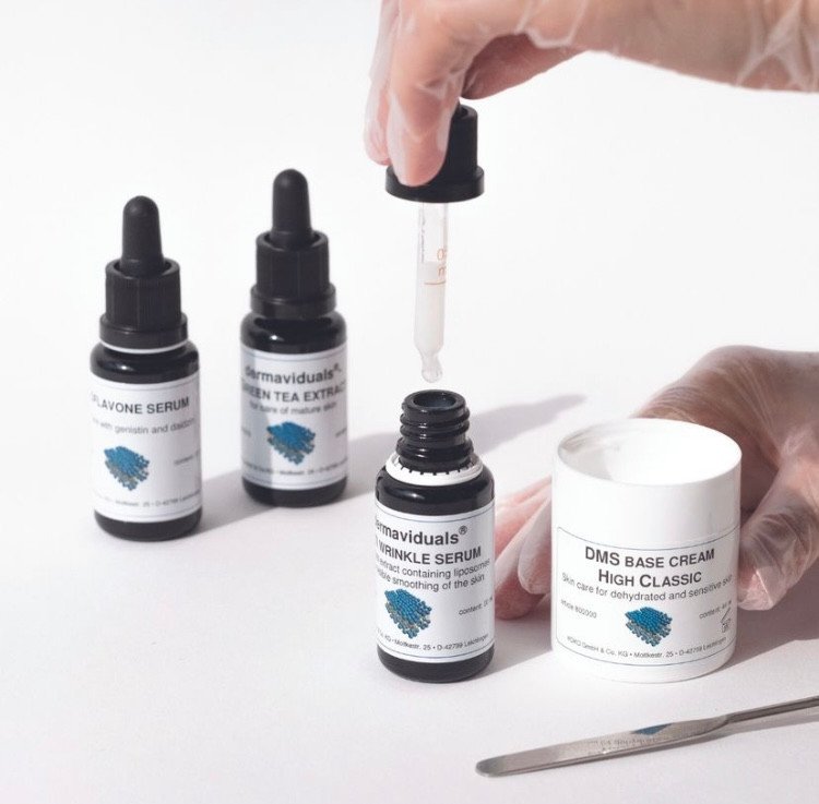Person wearing gloves using a dropper to transfer serum from a small bottle into a container, with other skincare products nearby.