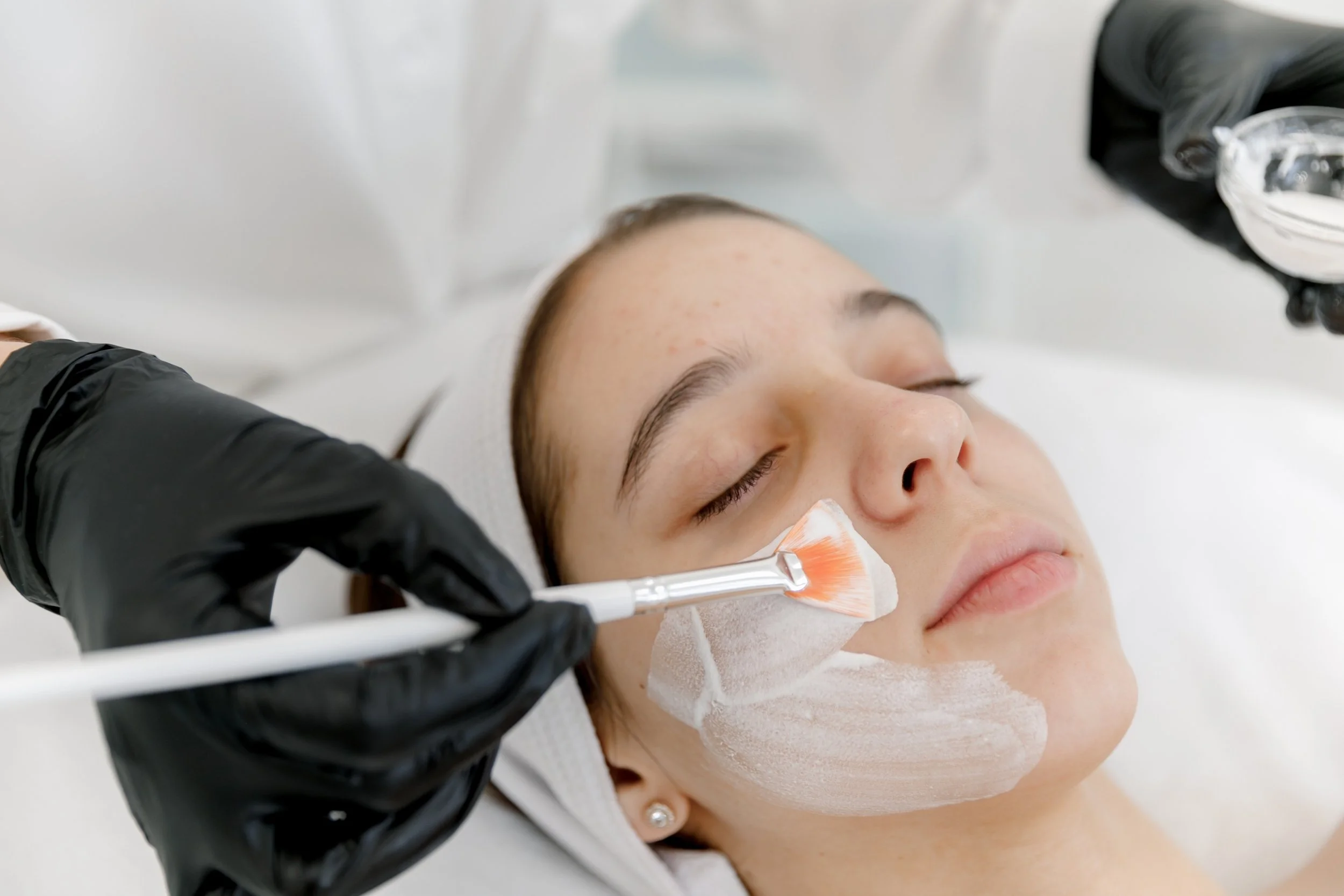 A woman with light skin and brown hair is lying down with her eyes closed while a person wearing black gloves applies a facial mask with a cosmetic brush.
