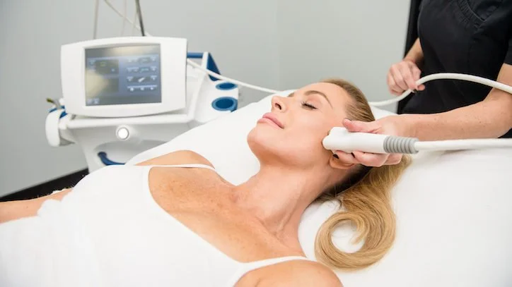 A woman receiving a cosmetic or dermatological treatment with a handheld device, lying on a medical bed in a clinical setting.