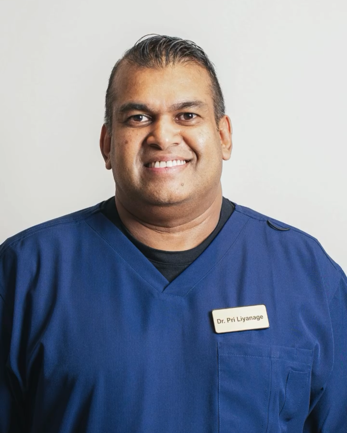 A smiling man in blue medical scrubs with a name tag that reads 'Dr. Pri Liyanage' against a plain light background.