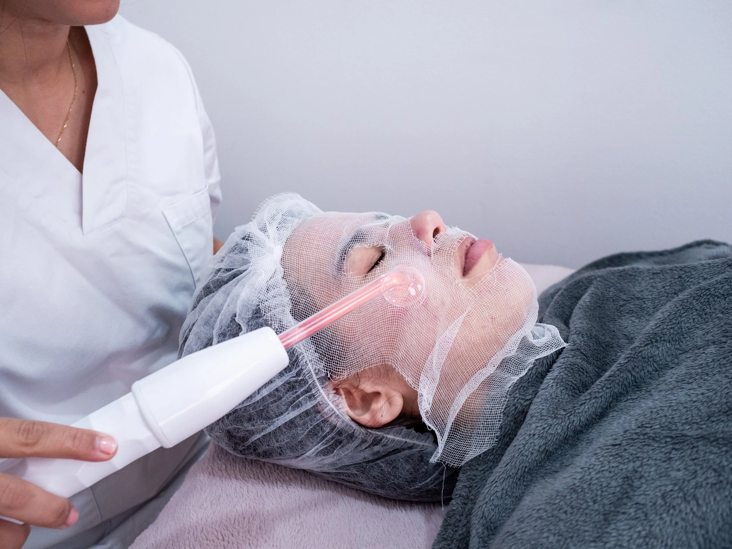 A person lying on a medical bed with a protective mesh covering their face, receiving a treatment from a healthcare professional in a clinical setting.