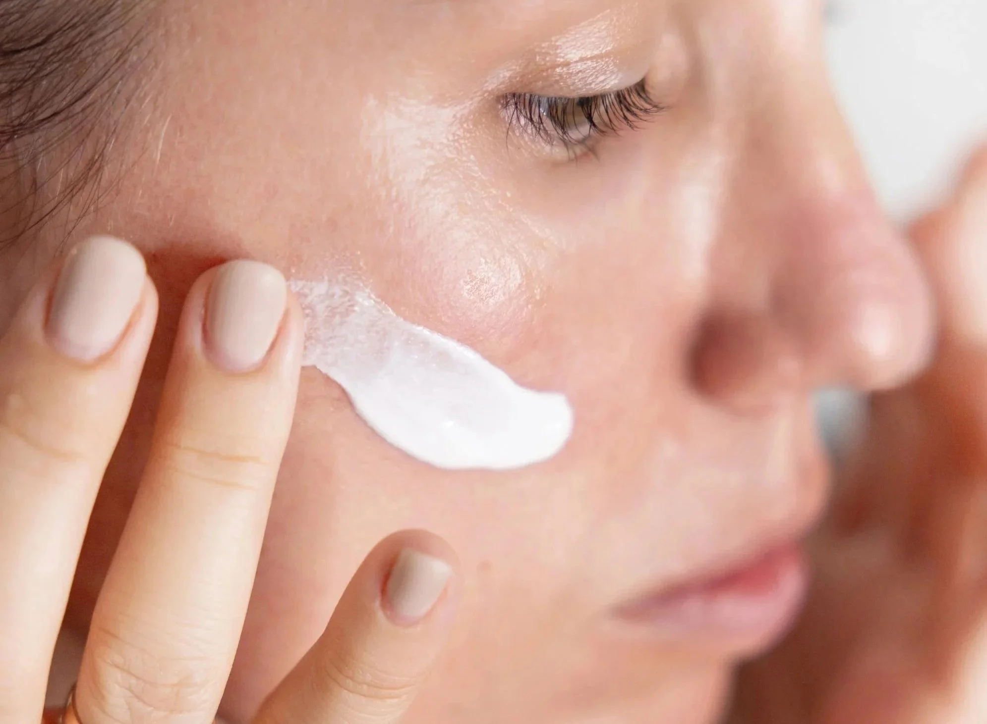 Close-up of a person applying skincare cream or moisturizer to their face, focusing on their cheek and eye area with fingers gently spreading the product.