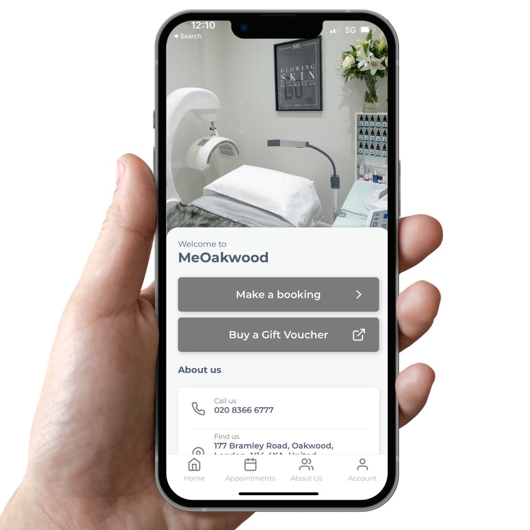 A smartphone screen showing the MeOakwood spa booking app with an interior image of a treatment room, featuring a white medical bed, a mounted piece of equipment, shelves with bottles, and a framed poster on the wall, held in a person's hand.