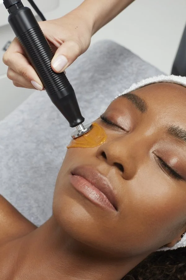 A woman receiving a cosmetic facial treatment with a handheld device, applying a golden mask on her cheek while lying down with her eyes closed.