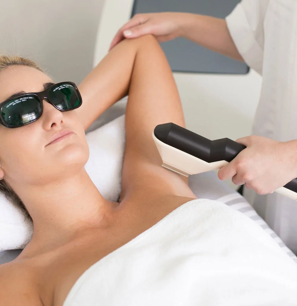 A woman receiving a laser hair removal treatment on her armpit while lying on a bed, wearing sunglasses, with a medical professional holding a laser device.