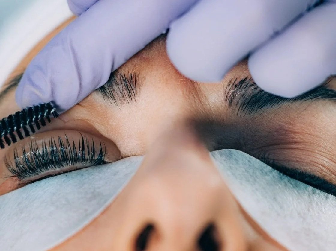 Close-up of a person receiving an eyelash extension treatment, with a technician applying the extension to the person's closed eye using a small brush and wearing gloves.