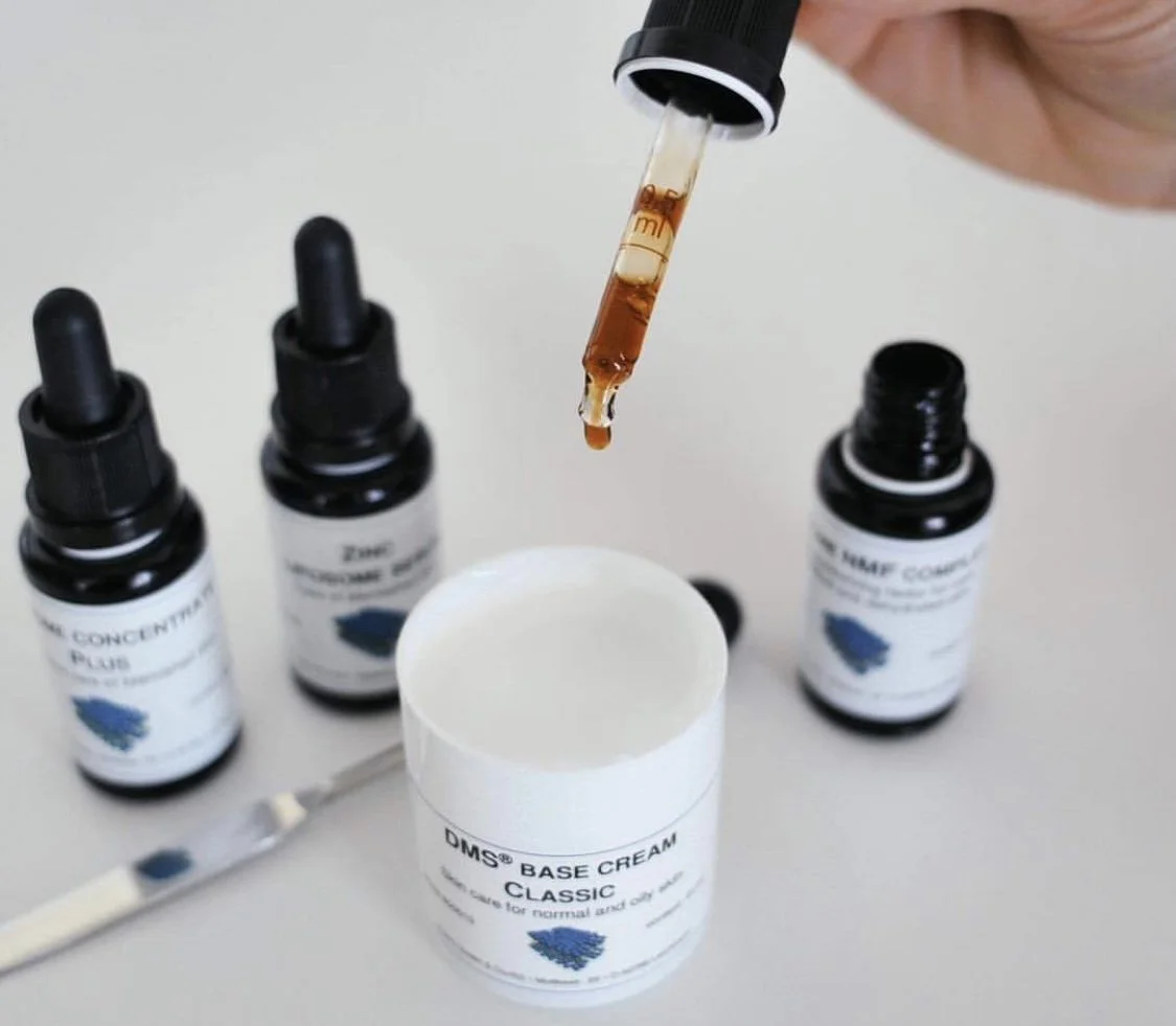 A skincare or cosmetic product setup with a dropper releasing a brown liquid into a white container labeled "DMS Base Cream" and three small bottles labeled "CONCENTRATE PLUS" and "ZOE" on a flat surface.