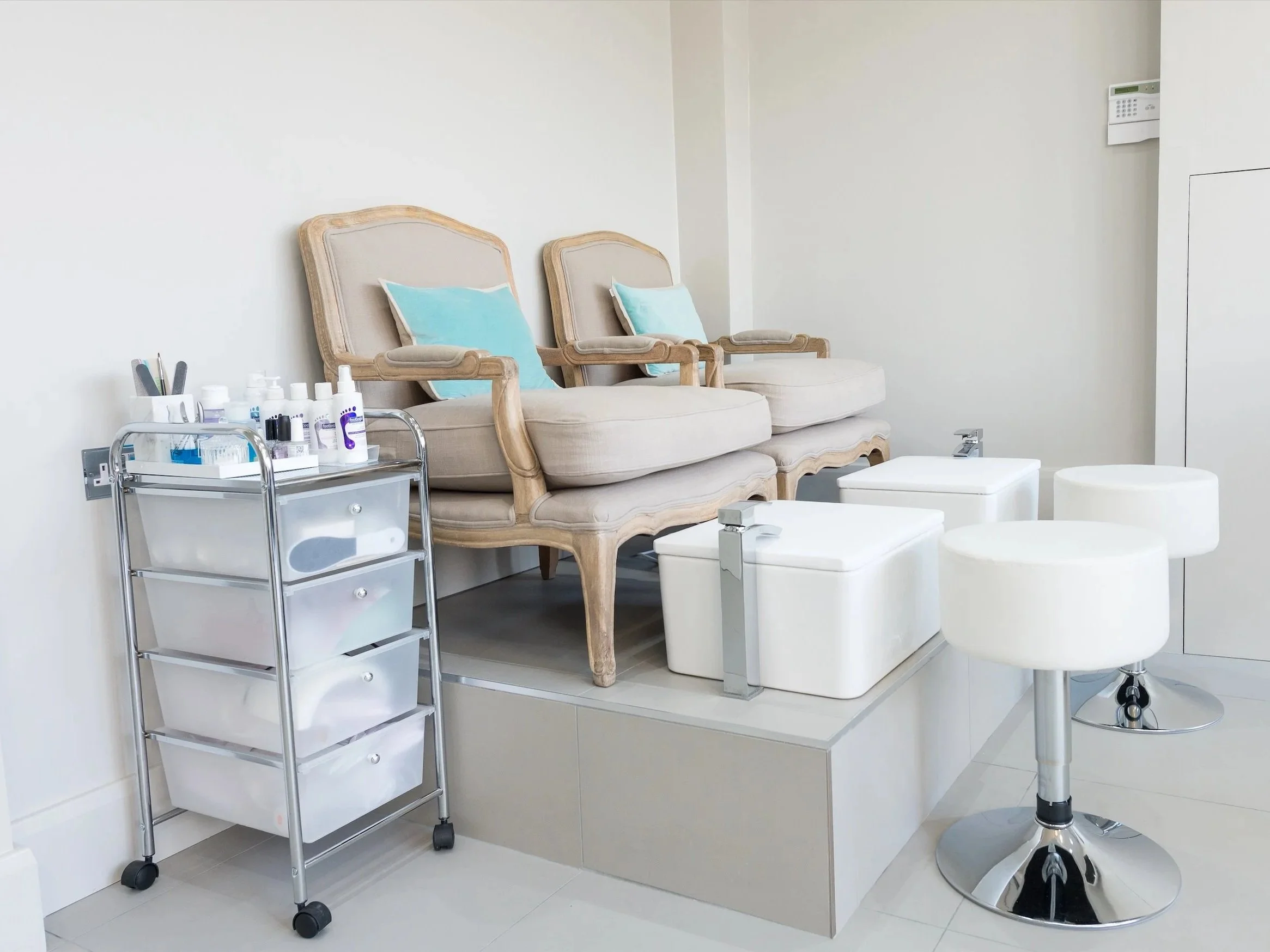 Medical treatment room with two beige vintage armchairs, each with a light blue pillow, a white side table with medical supplies, and a white stool on castors. The room has white walls and tile flooring.