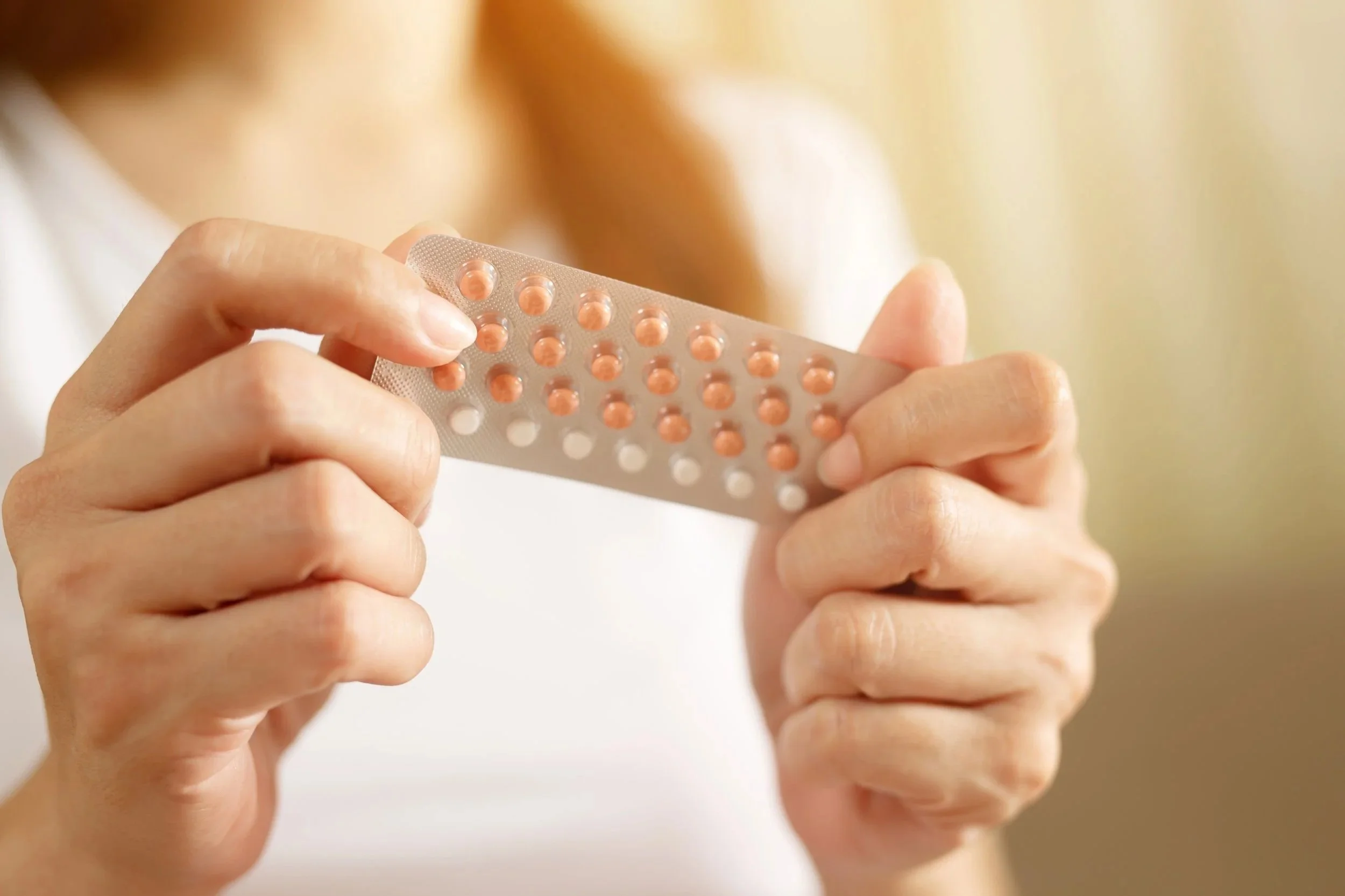 Close-up of a person holding a blister pack of round, peach-colored pills with some white pills, against a blurred background.