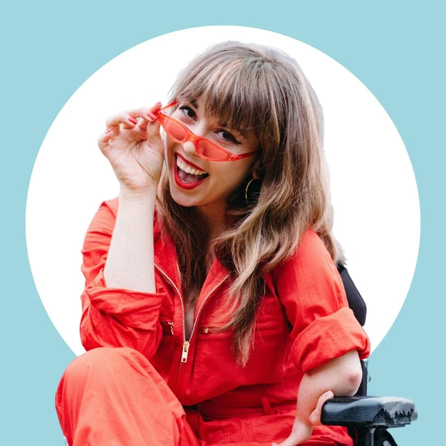 Young woman smiling while wearing red sunglasses and a red jumpsuit, sitting in a wheelchair with a light blue background and white circle behind her.