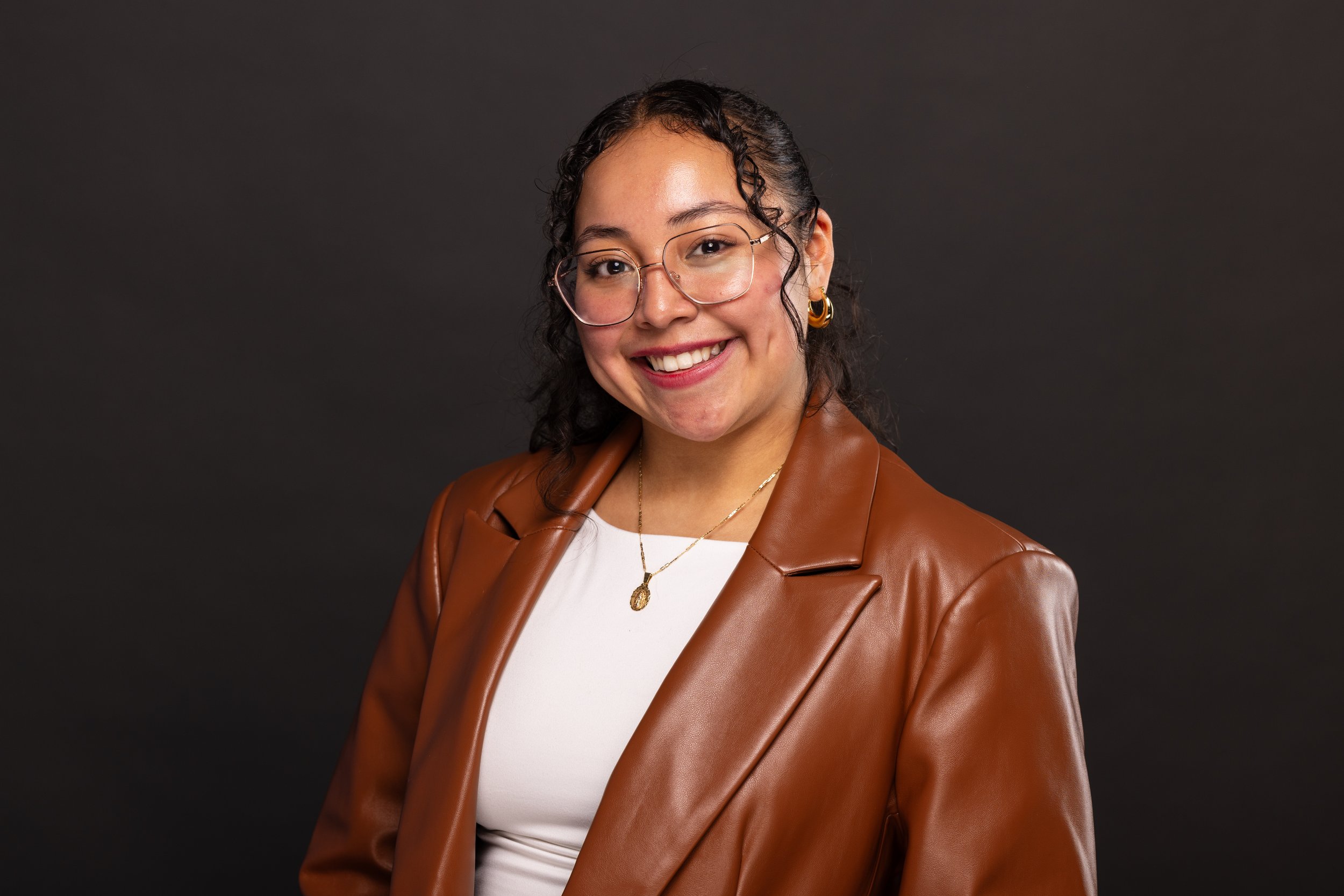 A woman smiling, wearing glasses, gold earrings, a brown leather jacket, a white shirt, and a gold necklace, standing against a dark background.