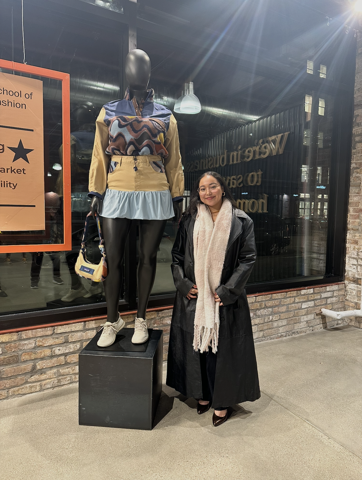 A woman standing next to a fashion mannequin display outside a store, both dressed in winter clothing. The woman is wearing a long black coat, a cream-colored scarf, and high heels. The mannequin is dressed in a colorful sweater, beige and blue layers, and is holding a small bag.