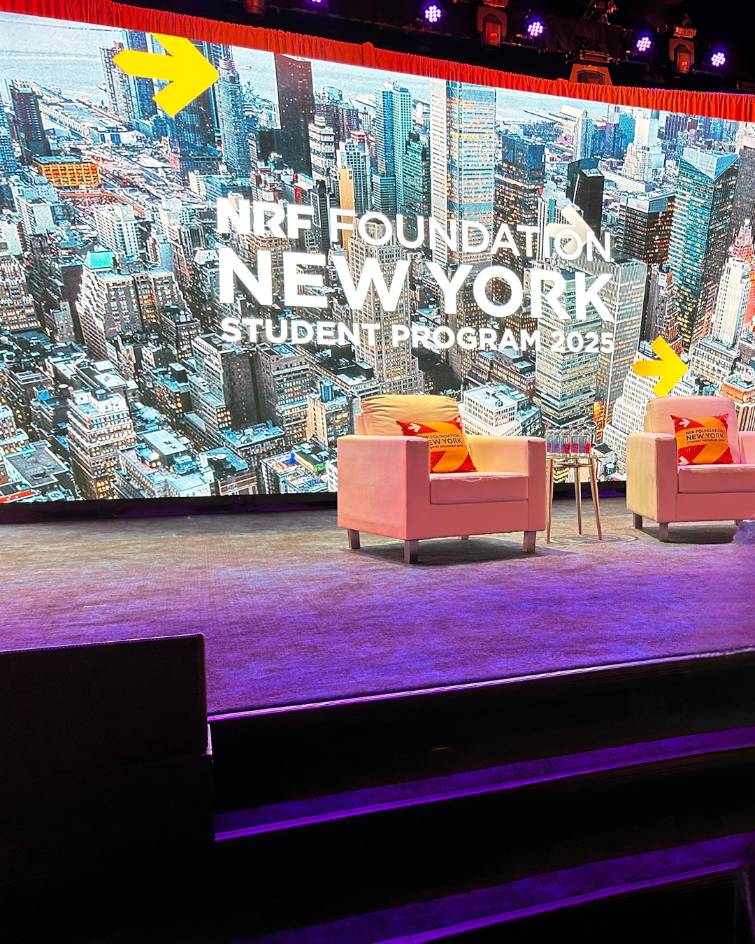 Stage with two pink chairs and a small table in front of a large screen displaying a cityscape of New York City with the text 'NRF Foundation New York Student Program 2025' and yellow arrows pointing to the top and right side of the cityscape.