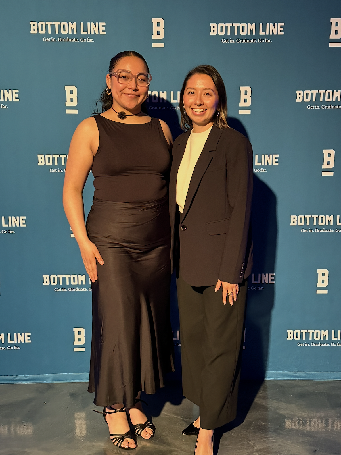 Two women standing on a stage in front of a blue backdrop with white text. One woman wears a black gown with a satin finish, glasses, and high heels, and the other wears a black suit with a cream blouse and black shoes. Both are smiling.