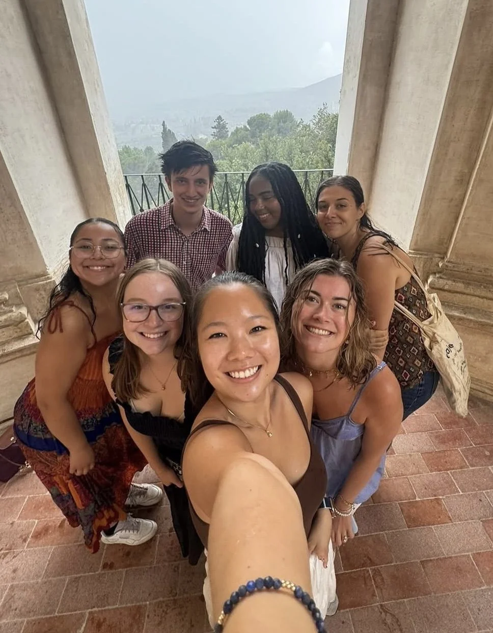 A group of seven young women and one young man taking a selfie together outdoors on a balcony with a scenic view of trees and mountains in the background.