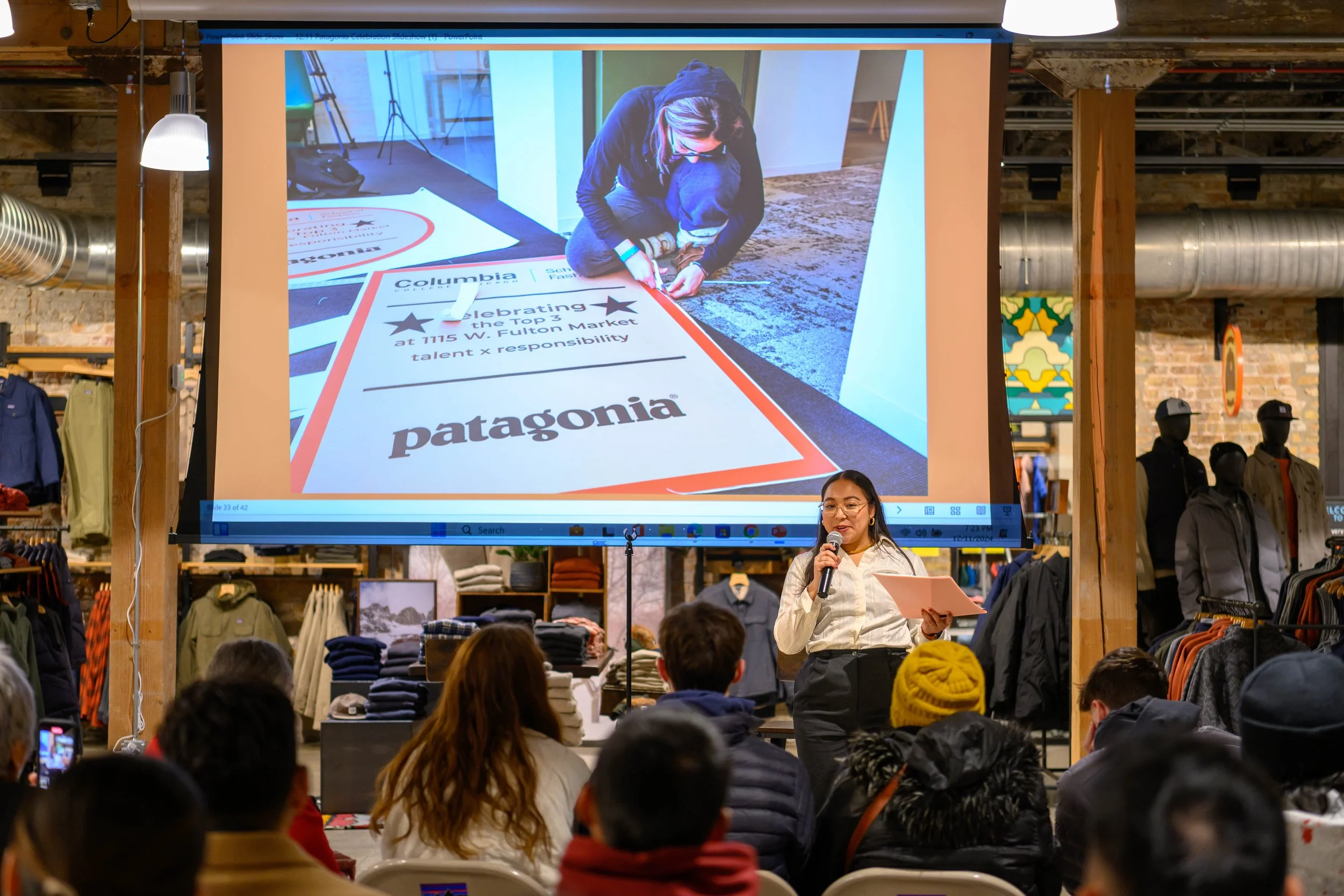 A woman is giving a presentation in a store, with a large screen behind her displaying a slide that has text about Columbia, Patagonia, and a store location. The store has clothing displays, mannequins, and people sitting in an audience.