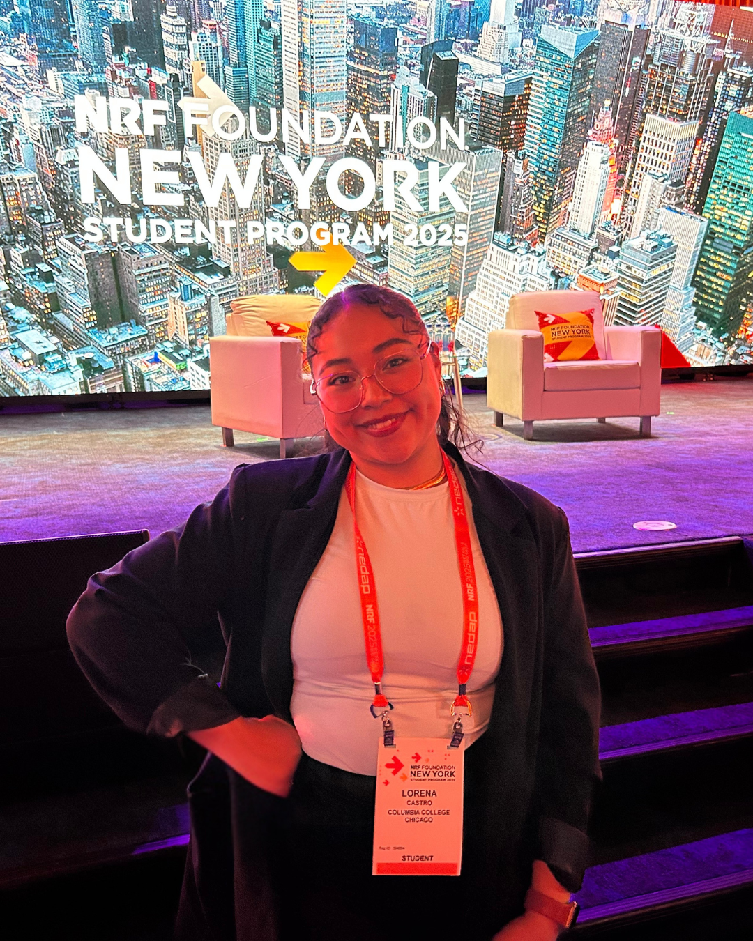 A young woman smiling at a conference stage with a lit cityscape backdrop. She is wearing glasses, a white shirt, and a black blazer, with an event pass around her neck that reads 'Lorena Castro, Columbia College, Chicago, Student.' The stage has two white chairs and the background displays I Heart NY Foundation on New York City skyline.