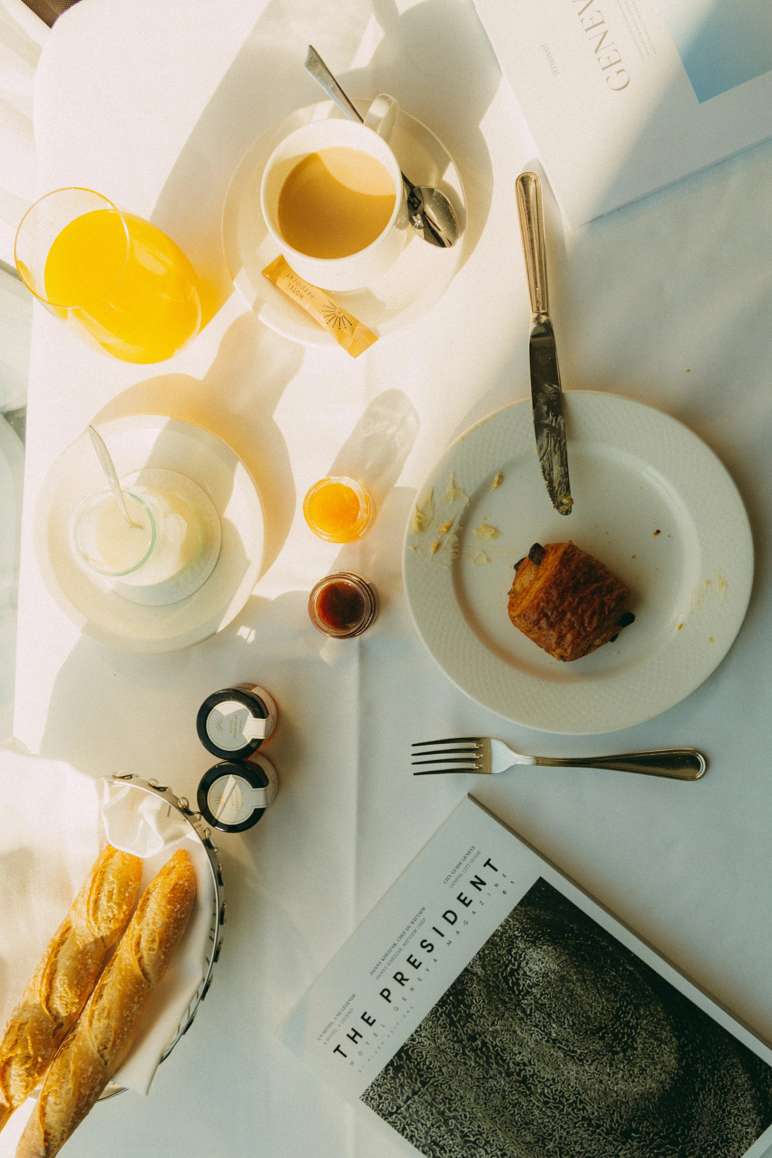 A breakfast table with a croissant, two glasses of orange juice, a cup of coffee, and some jars and condiments. There is a magazine and part of a menu on the table.