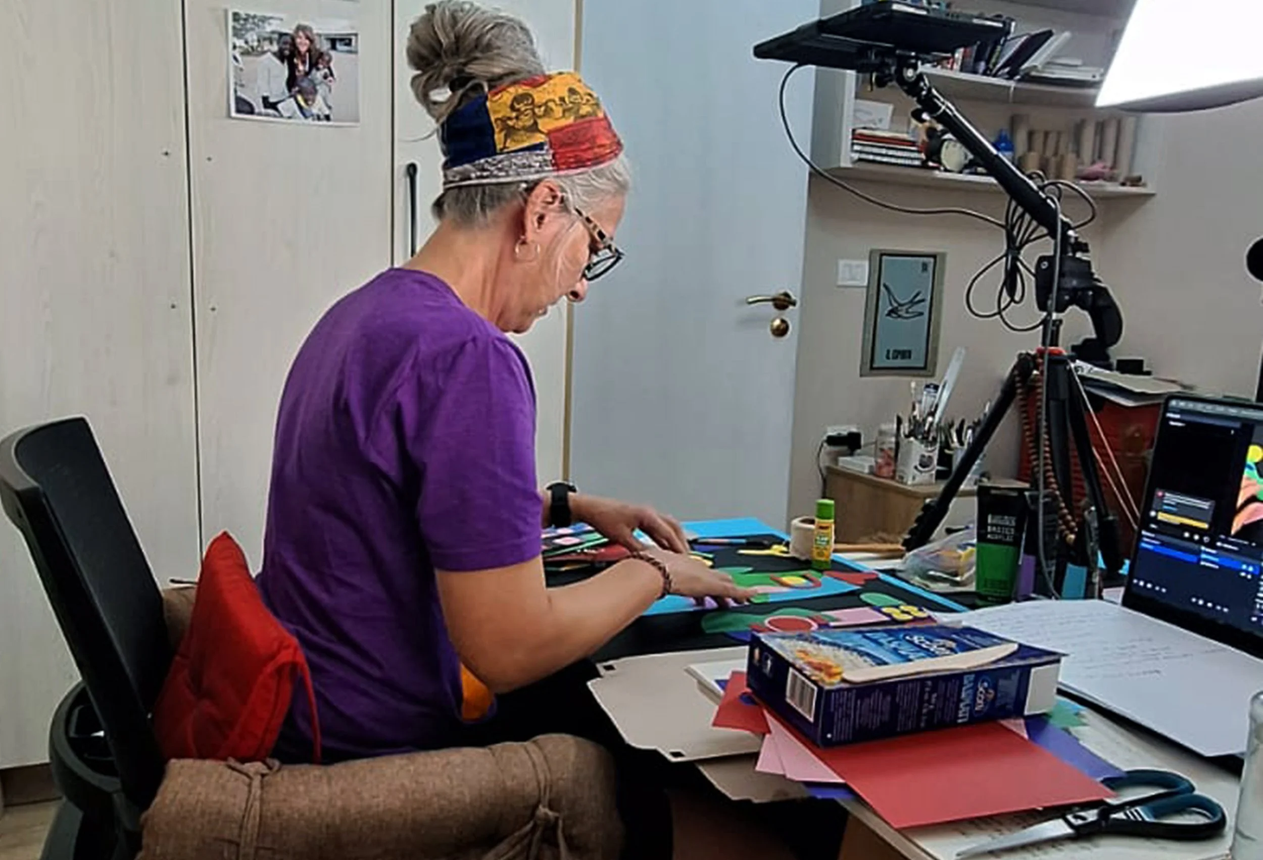 An elderly woman with glasses, gray hair tied up with a colorful headscarf, wearing a purple T-shirt, seated at a cluttered desk working on a colorful paper art project. The workspace includes glue, scissors, colored paper, a laptop, books, and a camera on a tripod.