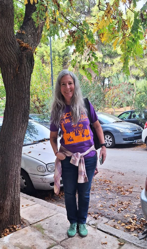 Smiling woman with long gray hair standing on sidewalk under a large tree, wearing a purple graphic T-shirt, dark jeans, teal sneakers, and a pink plaid shirt tied around her waist, with parked cars and trees in the background.