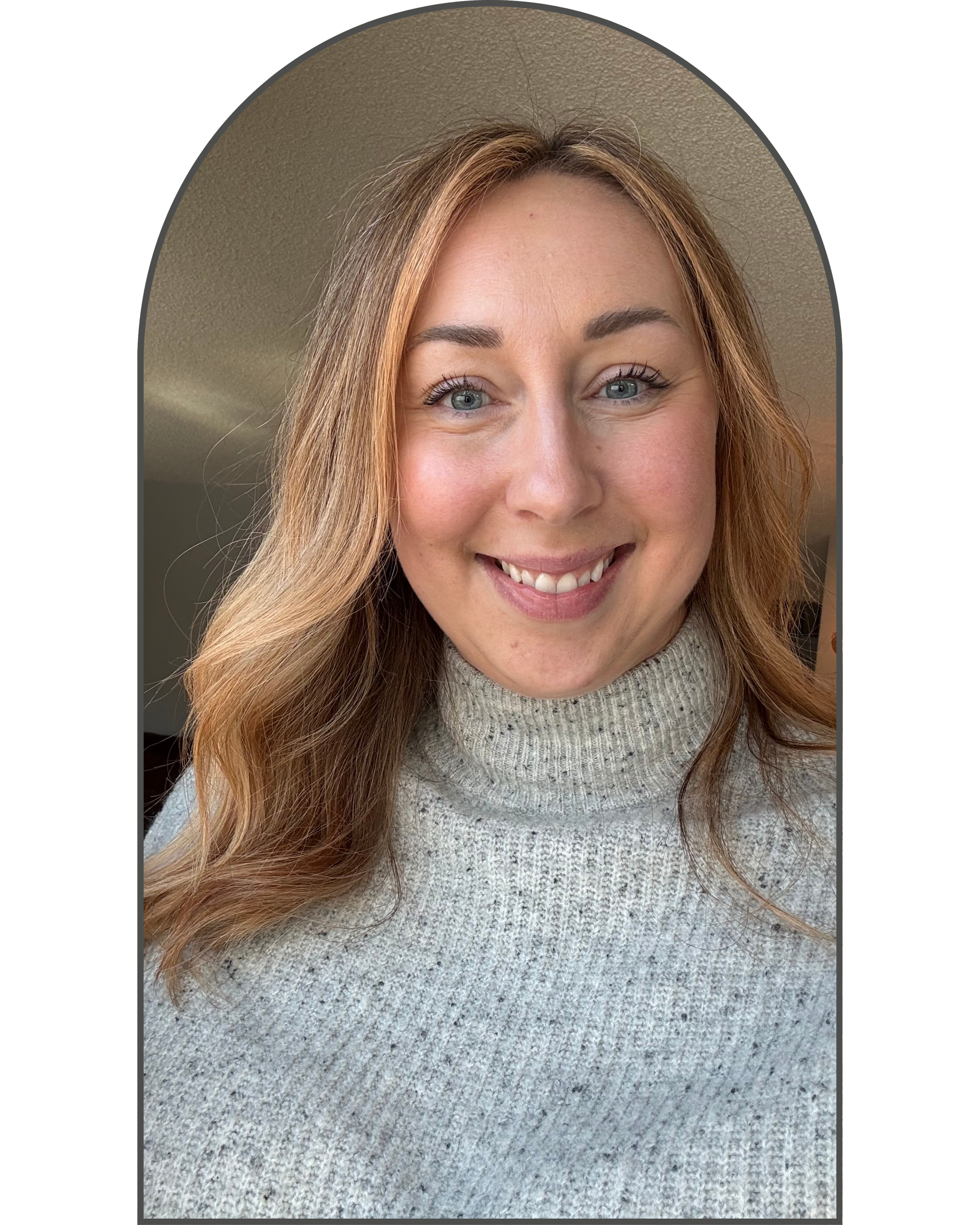 Close-up of a smiling woman with blonde hair and blue eyes, wearing a gray turtleneck sweater, indoors.