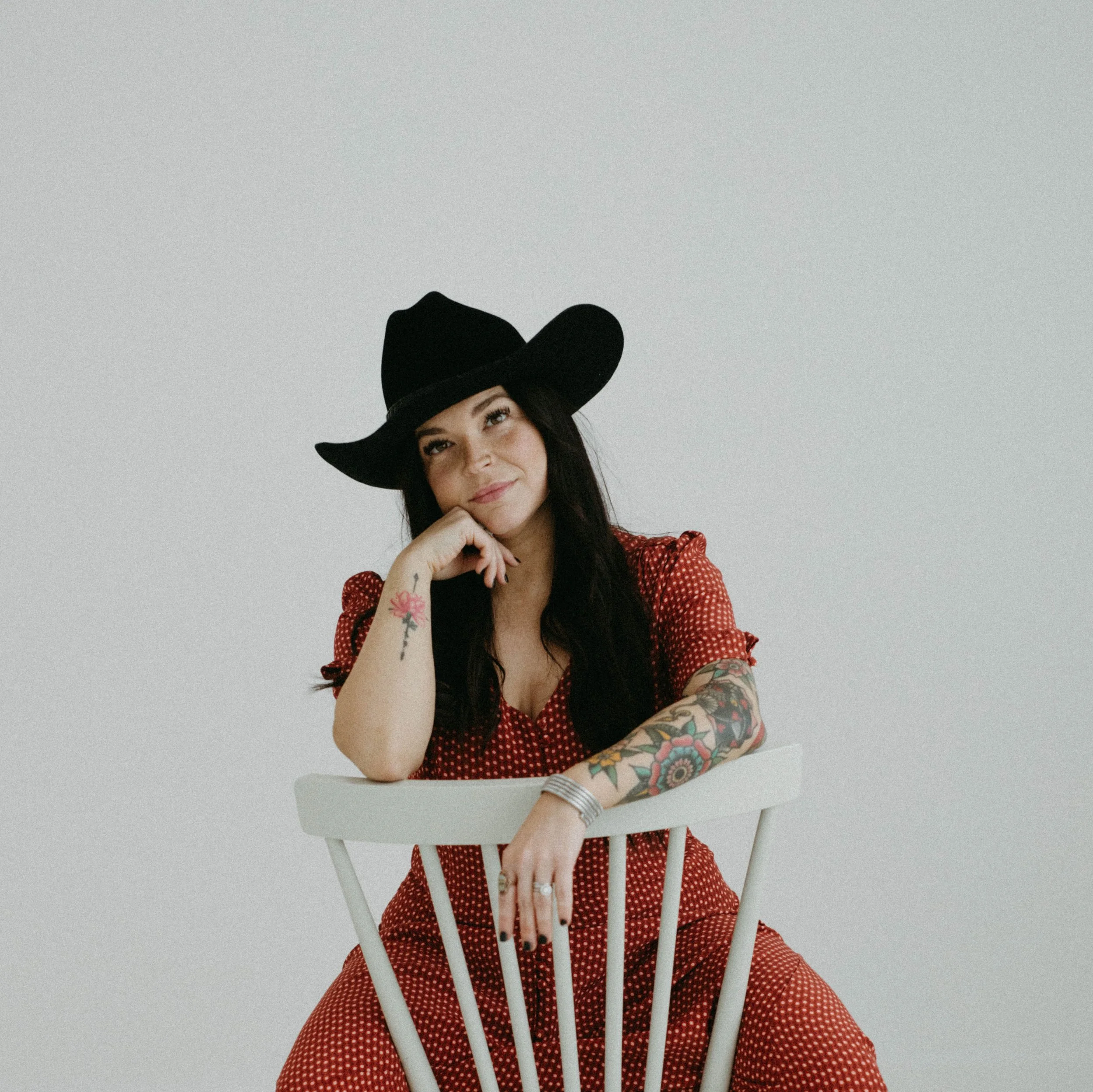 Woman with black hair and tattoos on her arms, wearing a black cowboy hat and a red polka dot dress, sitting on a white chair against a plain white background.
