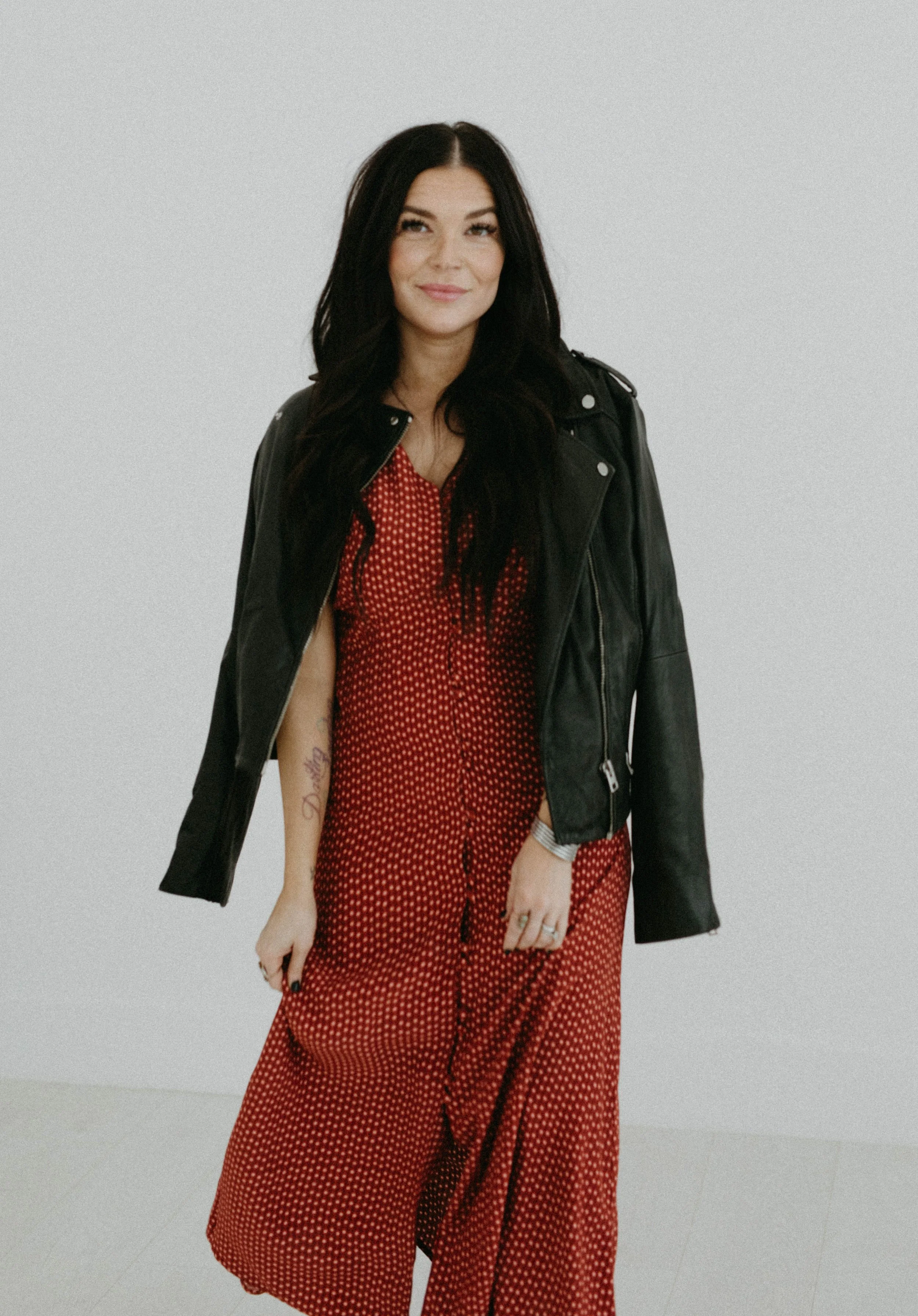 A woman with long dark hair wearing a red polka dot dress and a black leather jacket, standing against a plain white background.