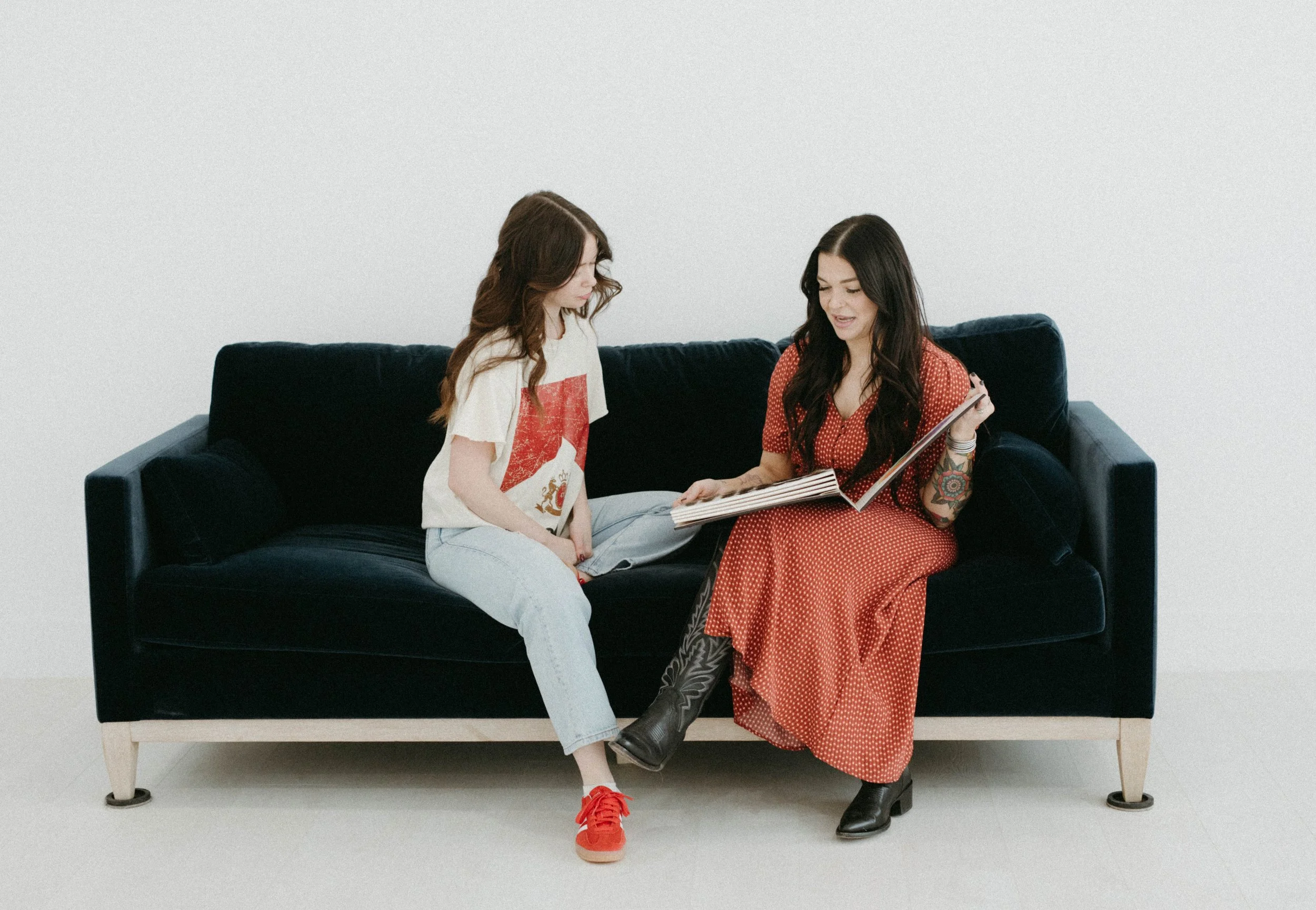Two women sitting on a dark blue sofa, one showing a photo album to the other in a minimalist room.