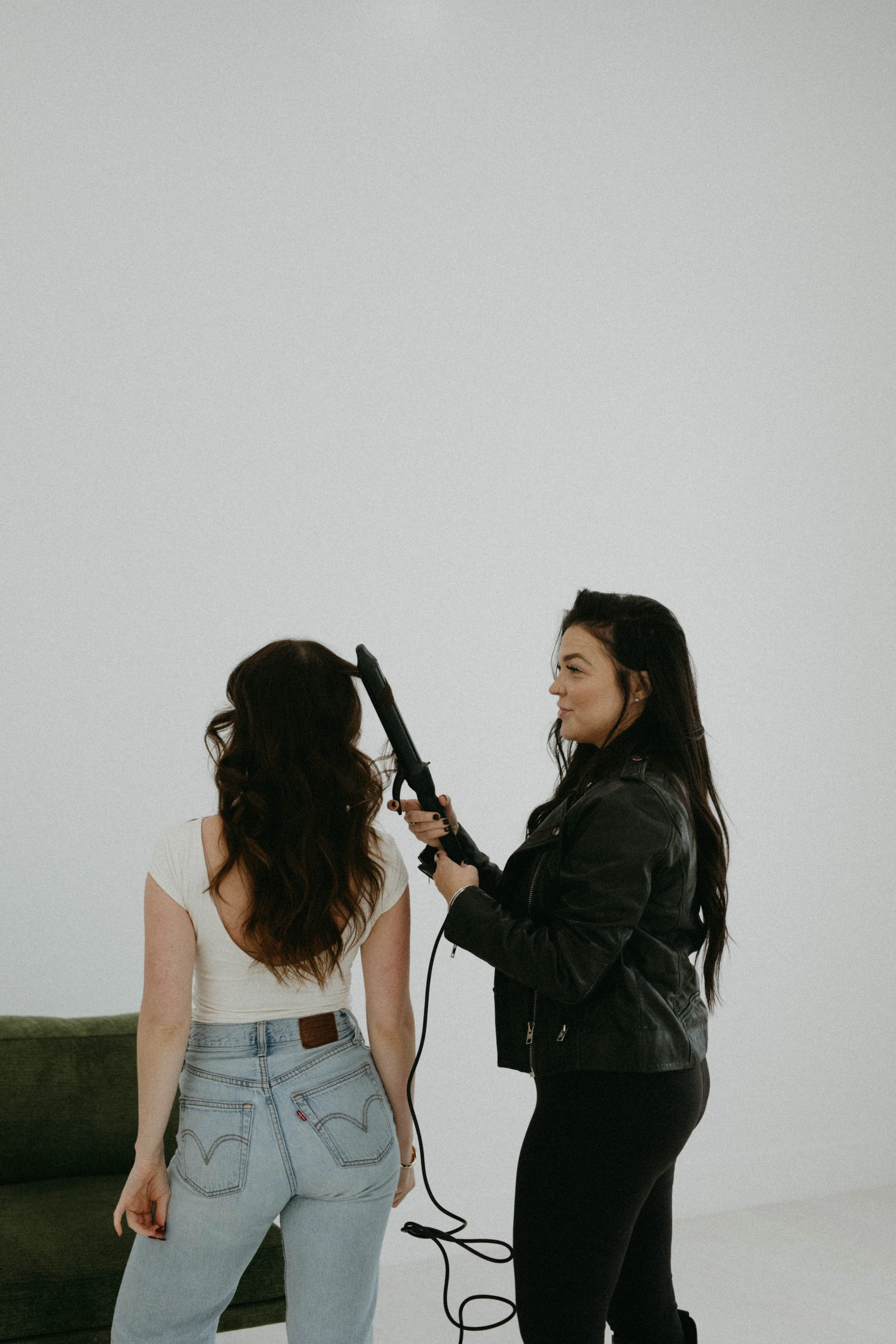 A woman styling another woman's hair with a curling iron in a minimalistic room.