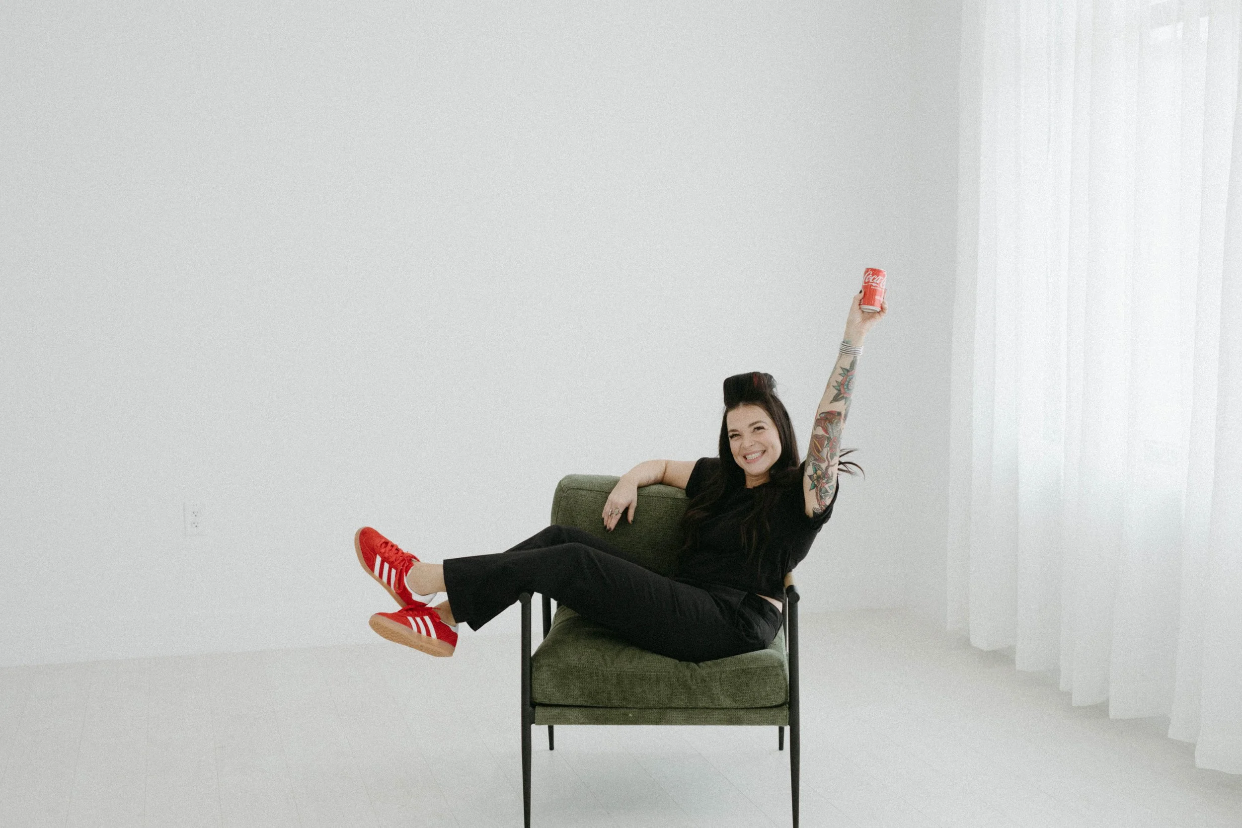 A woman with tattoos and red sneakers sitting on a green chair, smiling and raising a can of Coca-Cola in a minimalistic, white room.