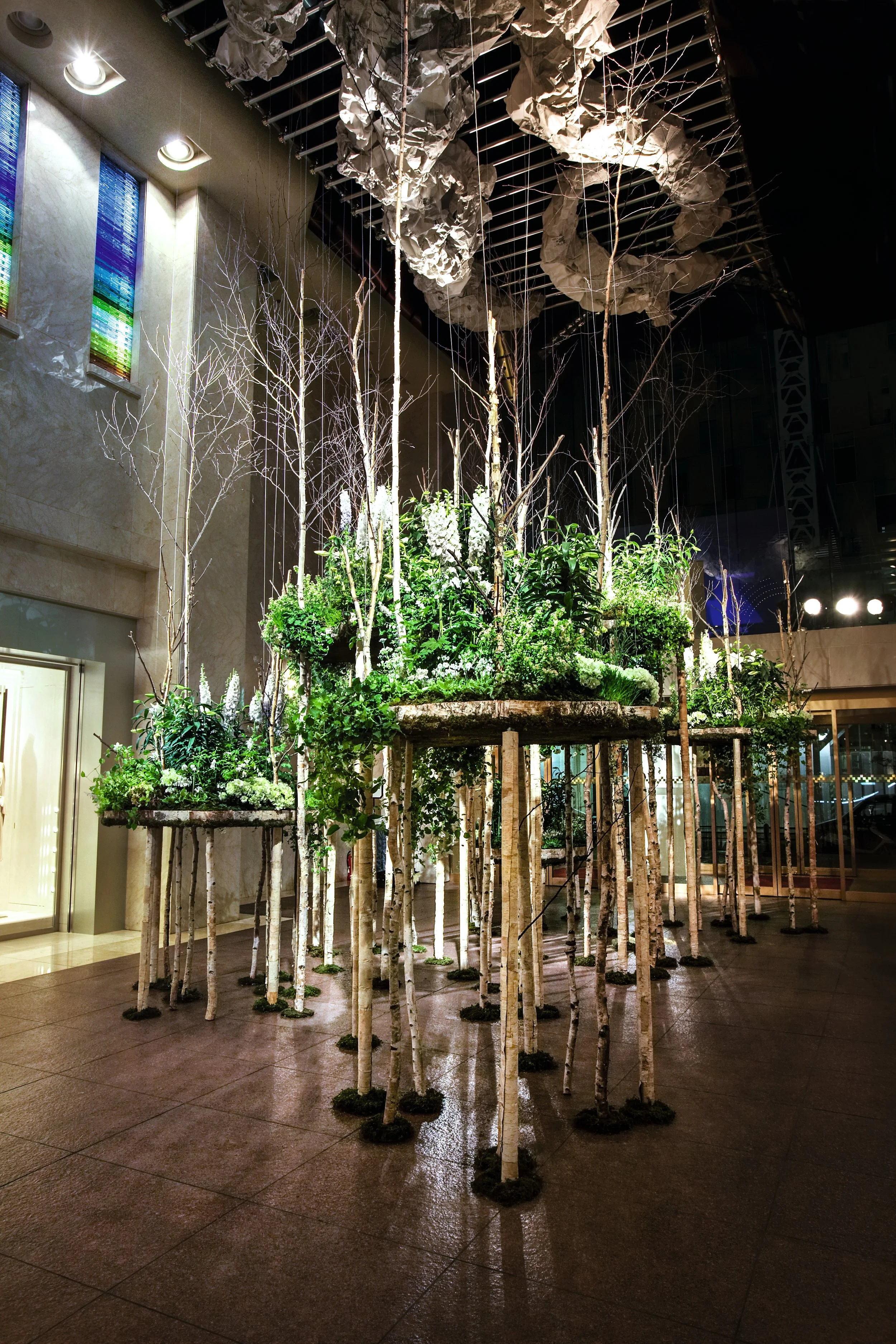 An indoor installation featuring tall, thin tree trunks and green plants on elevated platforms, with large crumpled paper-like structures hanging from a ceiling grid.