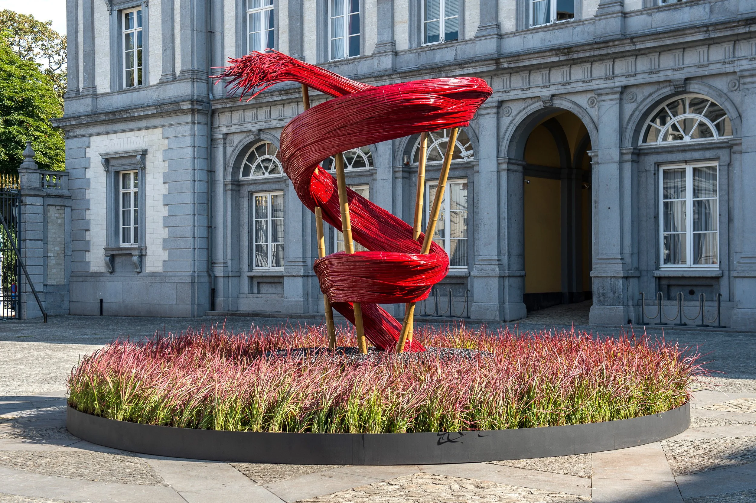 Sculptural floral installation with red woven spiral structure rising from ornamental grasses in front of a historic building facade in Brussels.