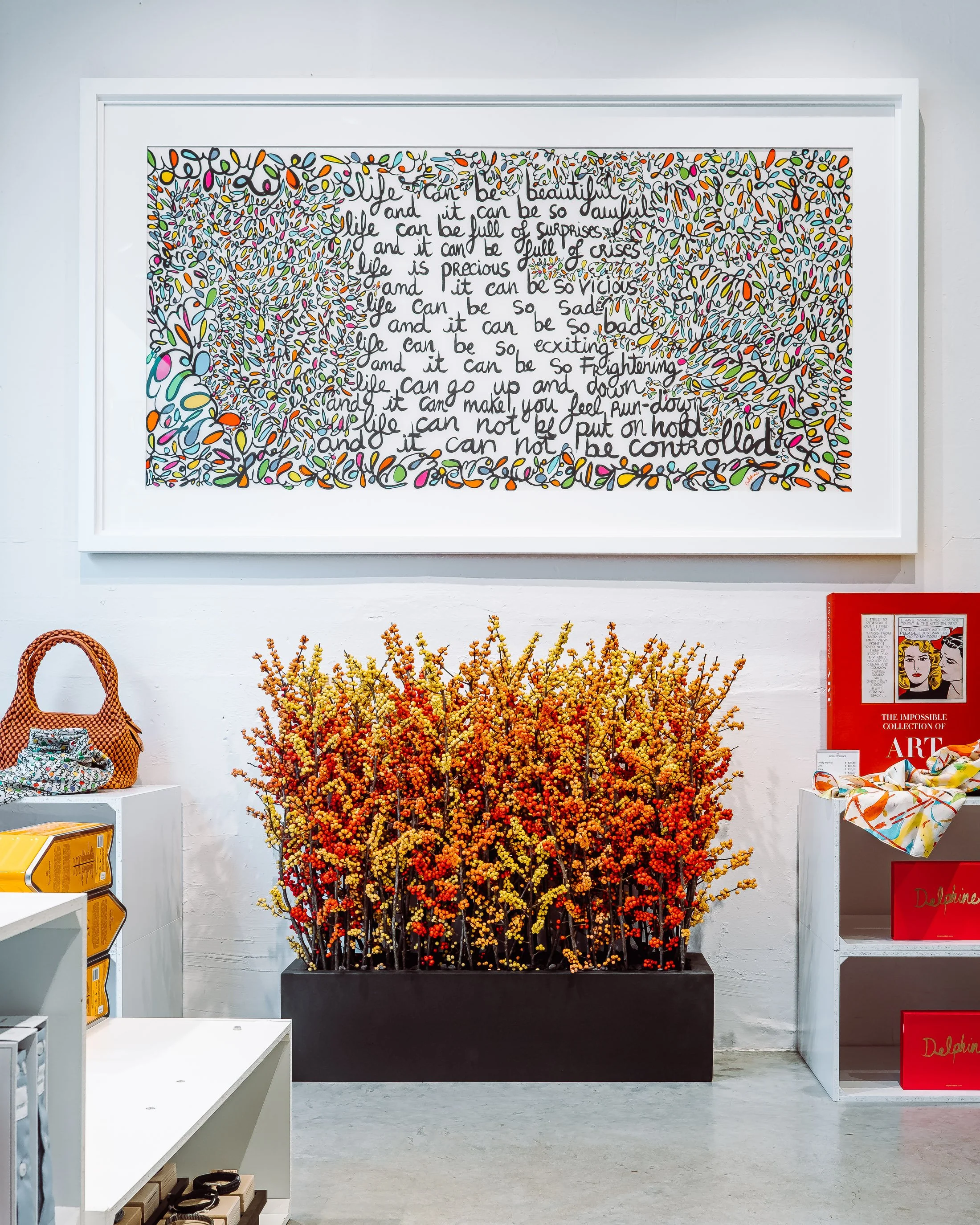 A colorful framed artwork with handwritten text and scribbles hangs on a white wall. Below, a large black planter contains orange and yellow blooming branches. To the right, a red display shelf with art books and a fabric piece, and on the left, a wi