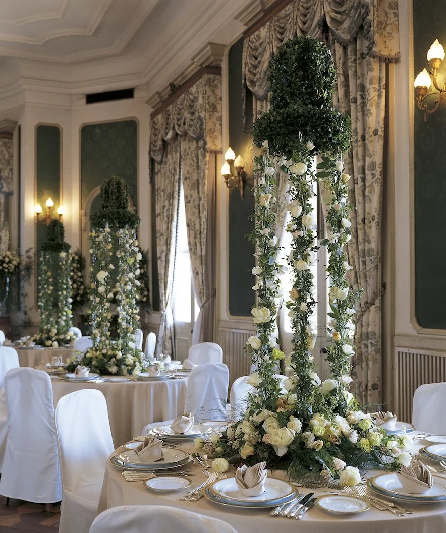 Round tables with tall green structures draped in white roses and ivy, evoking romance and intimacy in a classic wedding banquet hall.