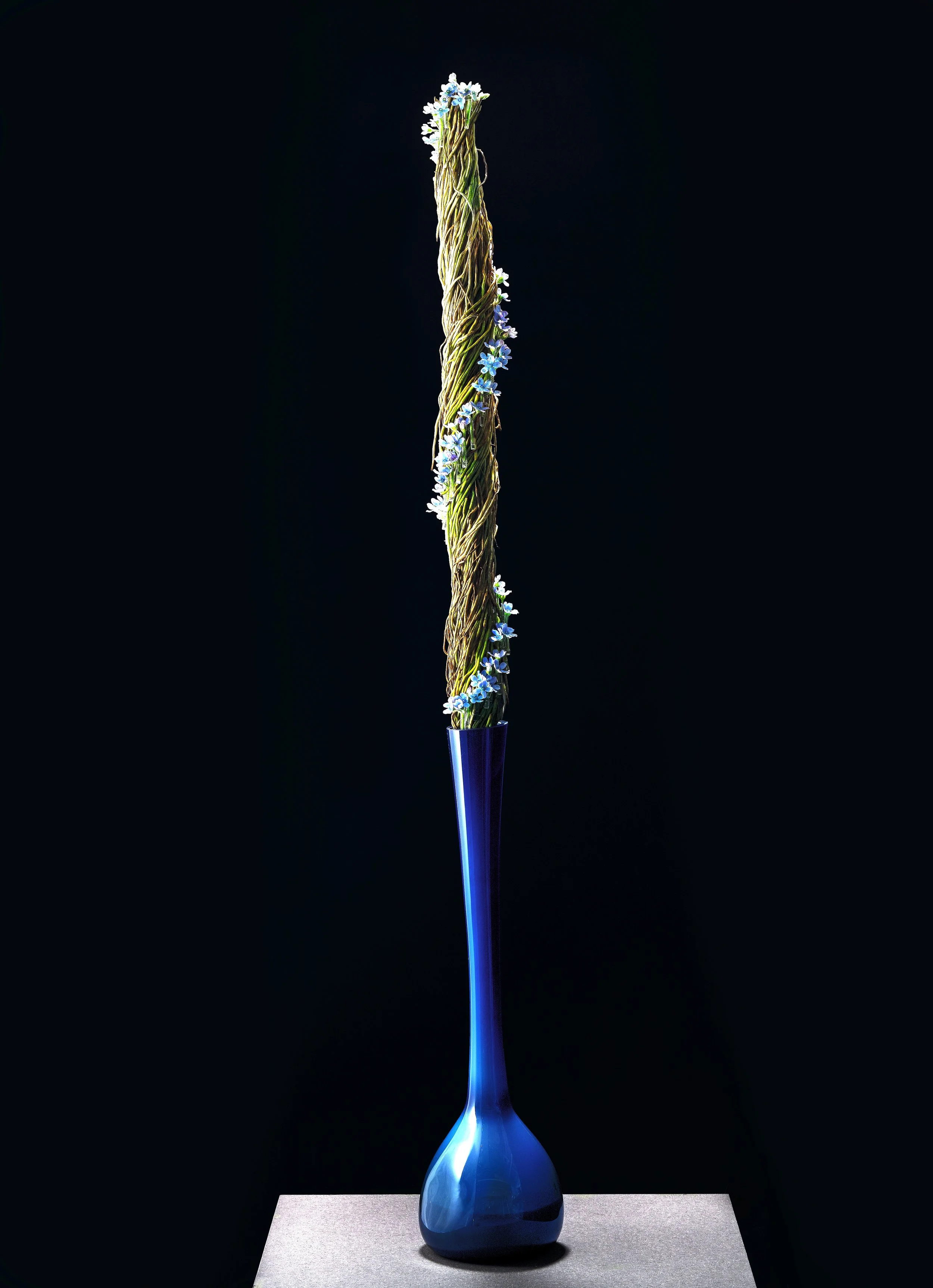 Tall blue glass vase holding a vertical twisted column of woven grasses adorned with delicate blue blossoms.