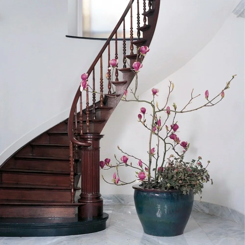 Daniel Ost magnolia floral arrangement in ceramic pot beside a wooden staircase.