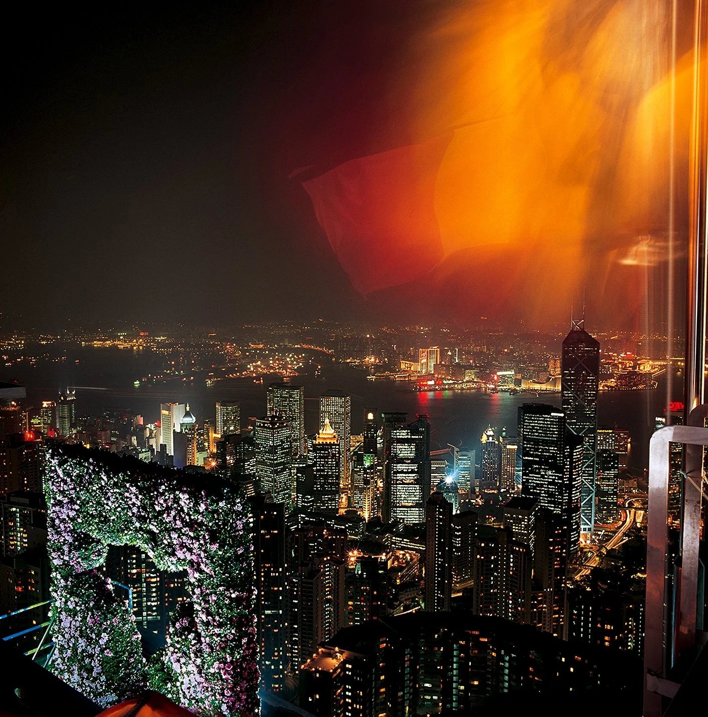 Hong Kong skyline at night with illuminated skyscrapers seen through a floral installation arch covered in pink blossoms.