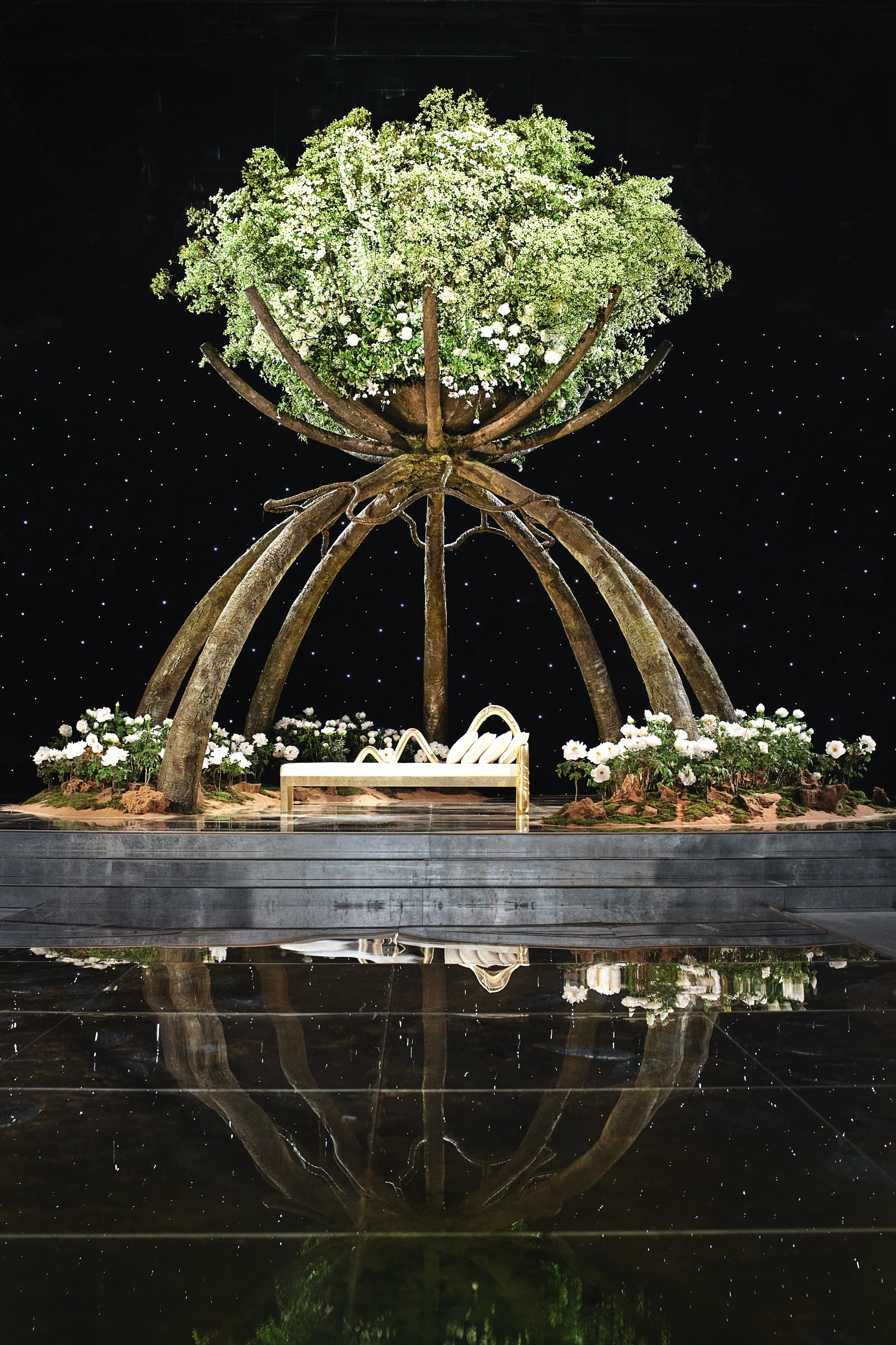 Wedding kosha stage with a giant sculptural floral tree canopy, white blossoms and greenery above a golden seating arrangement — an artistic centerpiece for a luxury celebration.