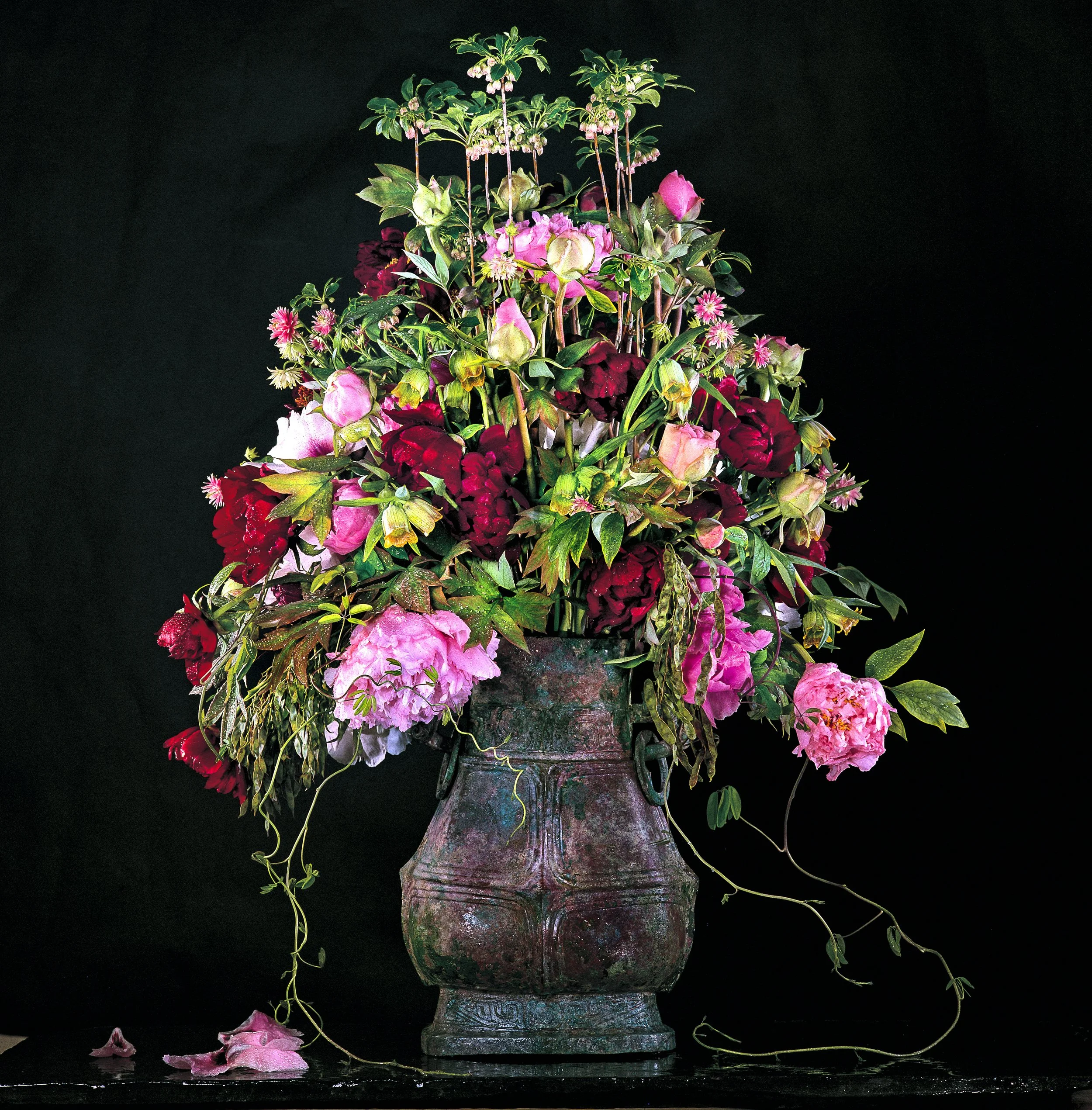 Romantic floral composition of peonies, roses, and celosia in shades of pink and burgundy, arranged in an antique bronze vase.