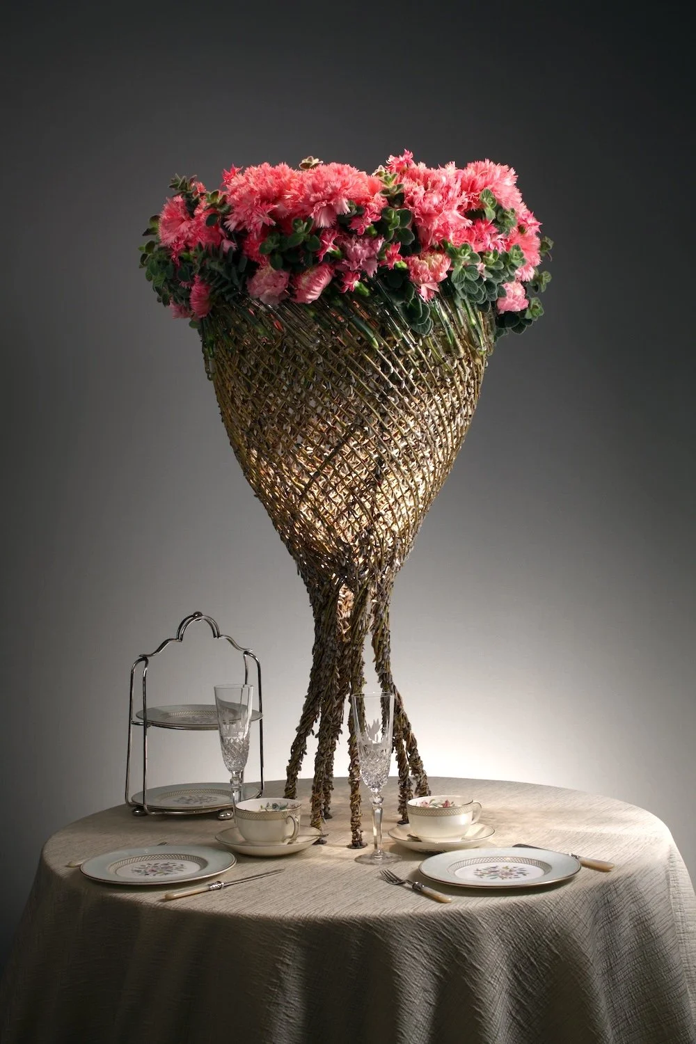 Artistic woven stand topped with vibrant pink flowers, forming a dramatic centerpiece for an intimate wedding reception table.