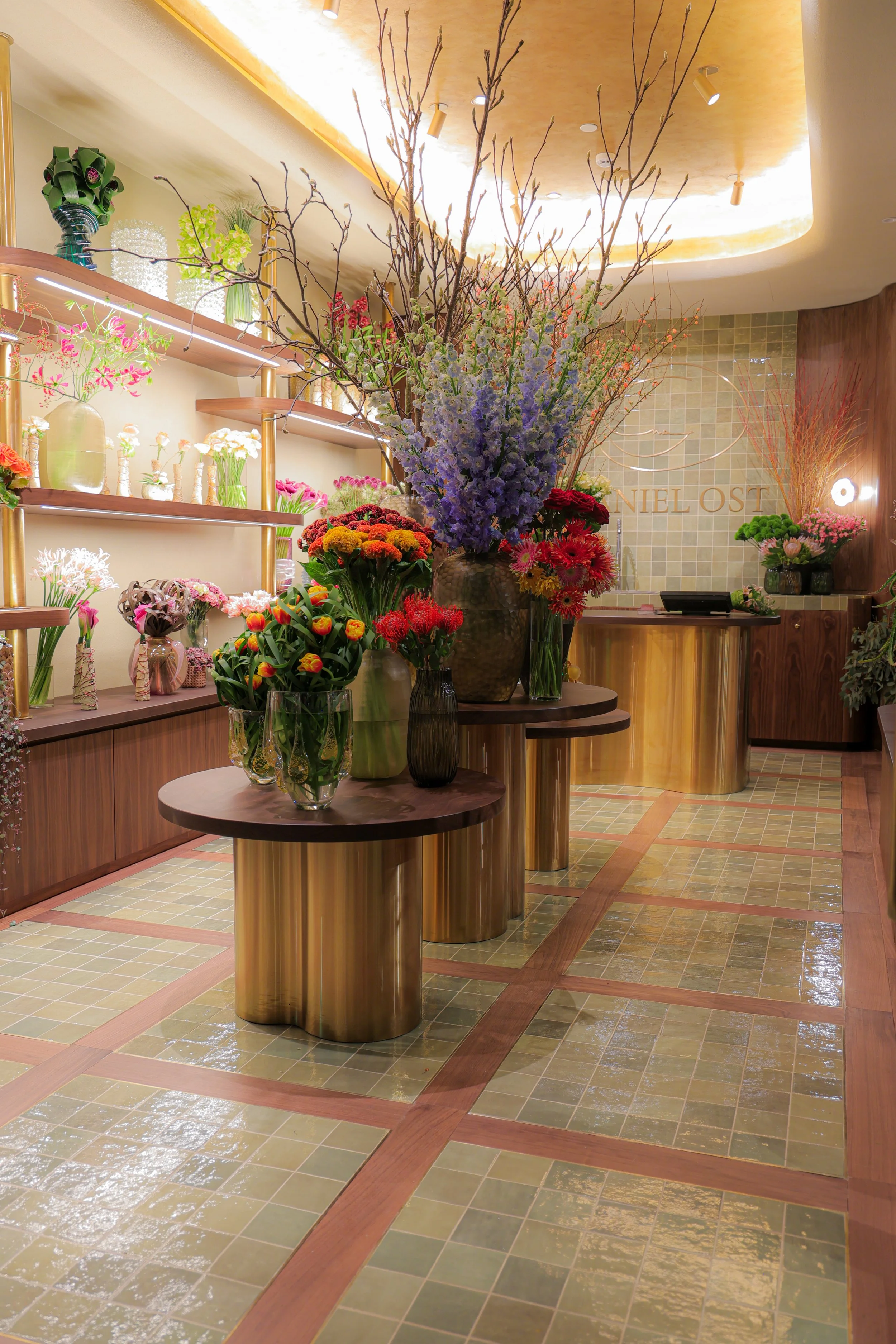 Interior of Daniel Ost flower shop in Riyadh with floral arrangements in vases on round tables and shelves, wooden accents, and warm lighting.