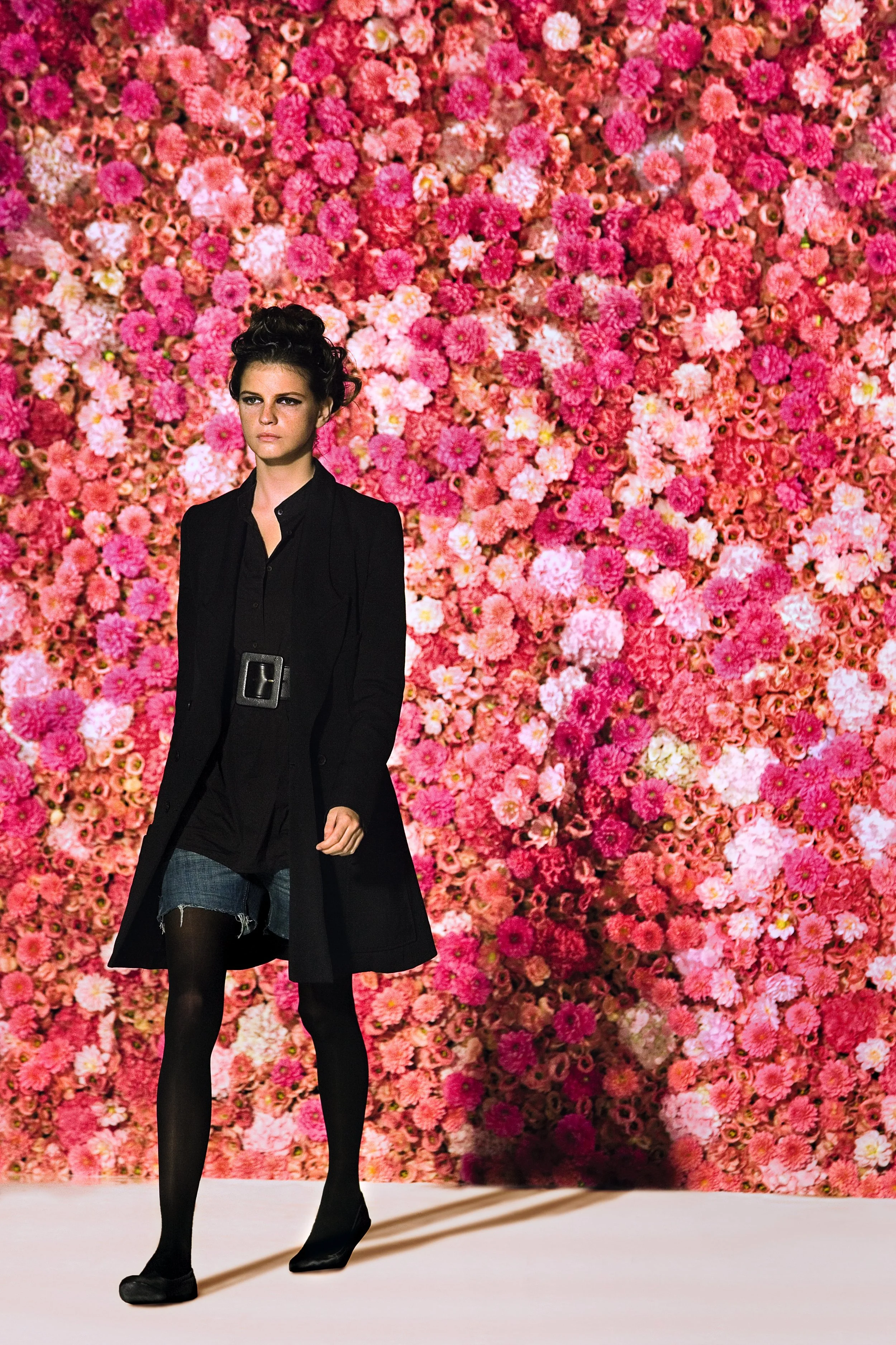 A fashion model walking on a runway in front of a background filled with pink and white flowers. The model is dressed in a black coat, black shirt, denim shorts, black tights, and black heels.