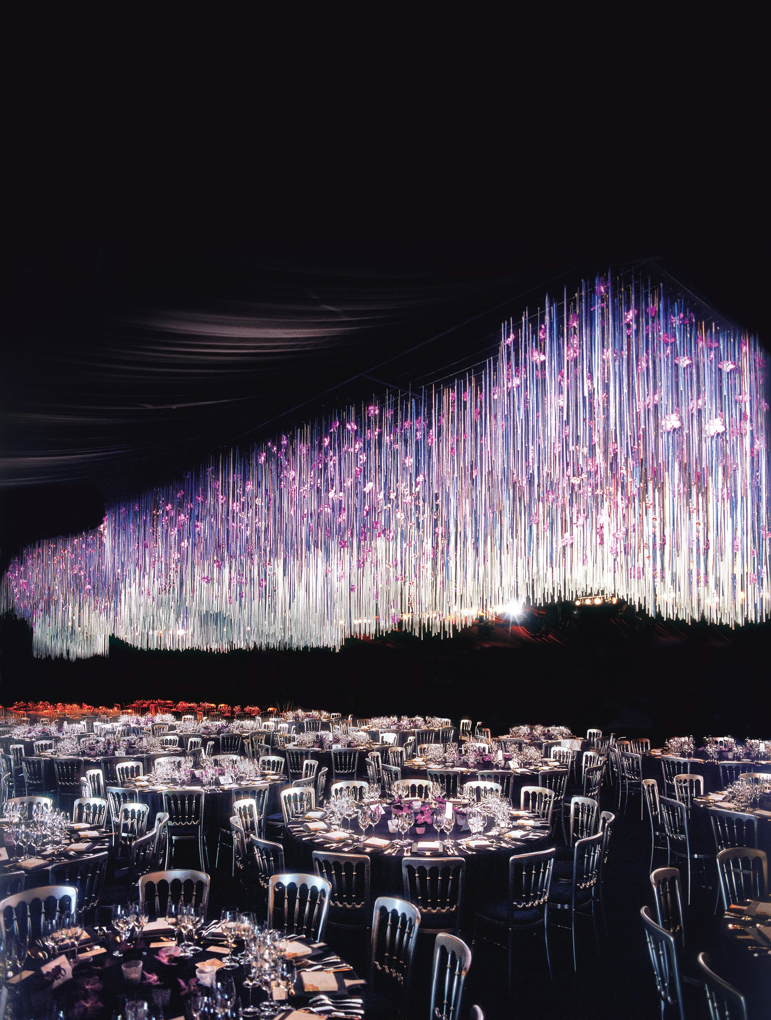 Corporate dinner with dramatic suspended installation of cascading violet and pink floral strands, atmospheric Daniel Ost design.