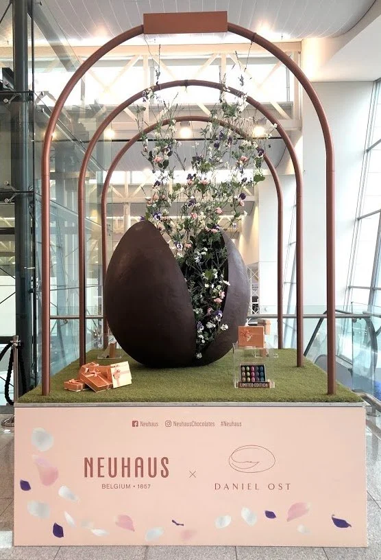 Neuhaus x Daniel Ost Easter installation with giant chocolate egg bursting with fresh flowers inside a luxury shopping mall display.