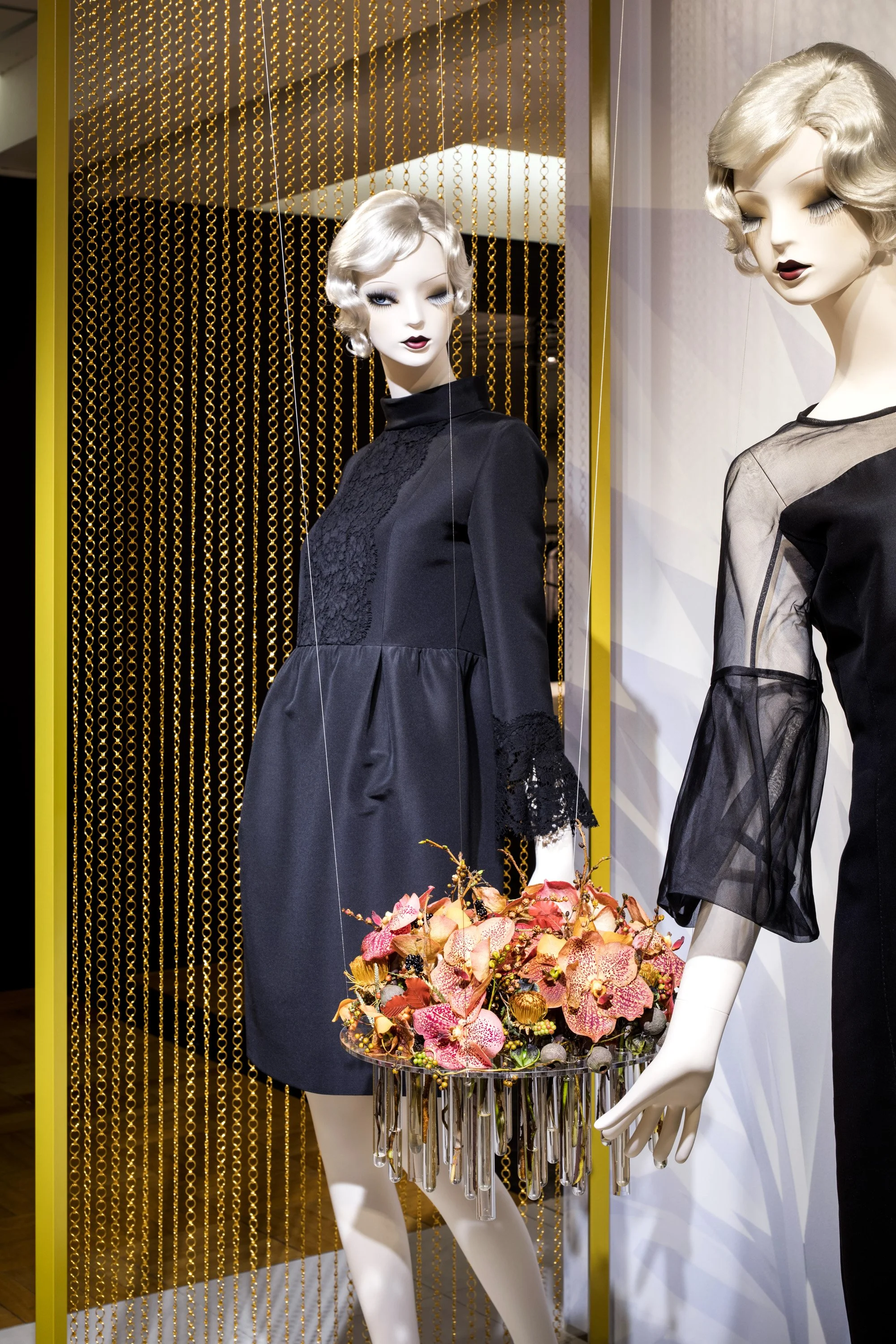 Store window display featuring two mannequins dressed in black, with a floral arrangement in a decorative bowl in front of them, and a gold chain curtain backdrop.