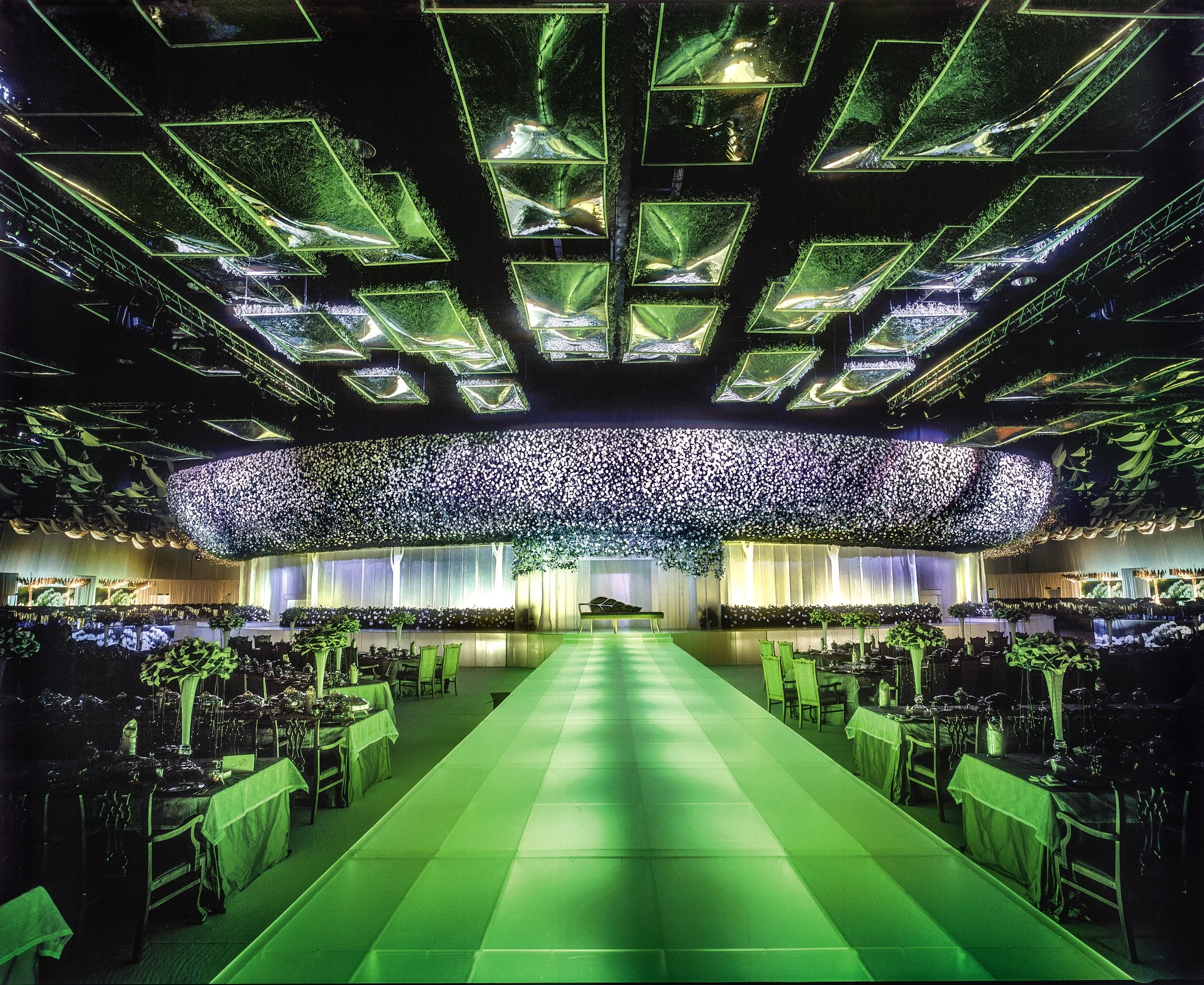 Grand wedding venue with dramatic green and white floral ceiling installation, mirrored panels, and illuminated green runway — modern luxury wedding decoration by Daniel Ost.