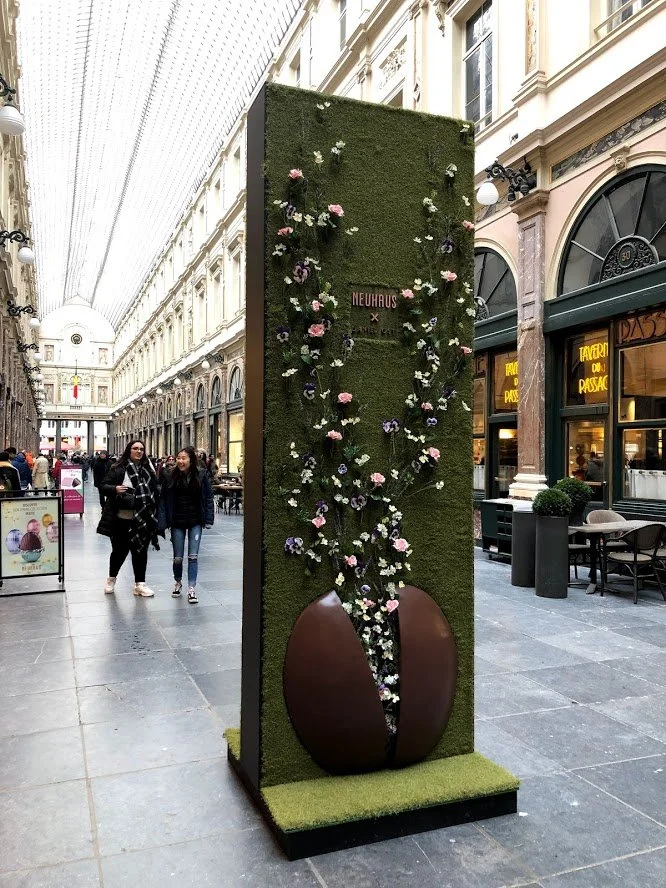 Public floral art installation in Galeries Royales Saint-Hubert, with vertical green wall and chocolate bean sculpture bursting with pastel flowers.
