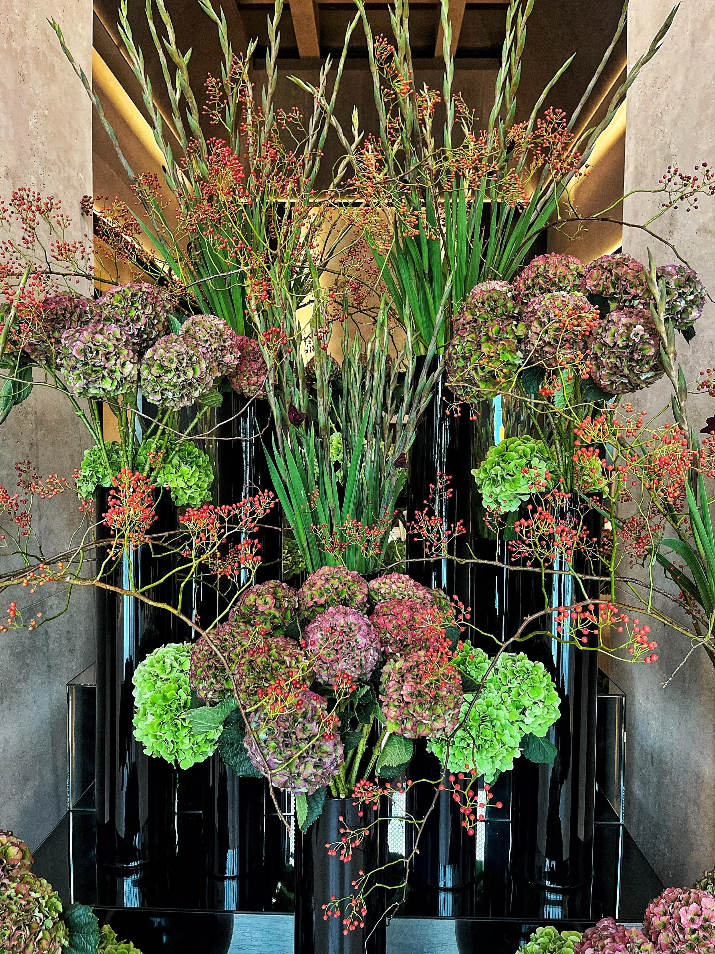Sculptural floral arrangement of hydrangeas, gladiolus, and berries in tall black vases for a hotel lobby interior design.